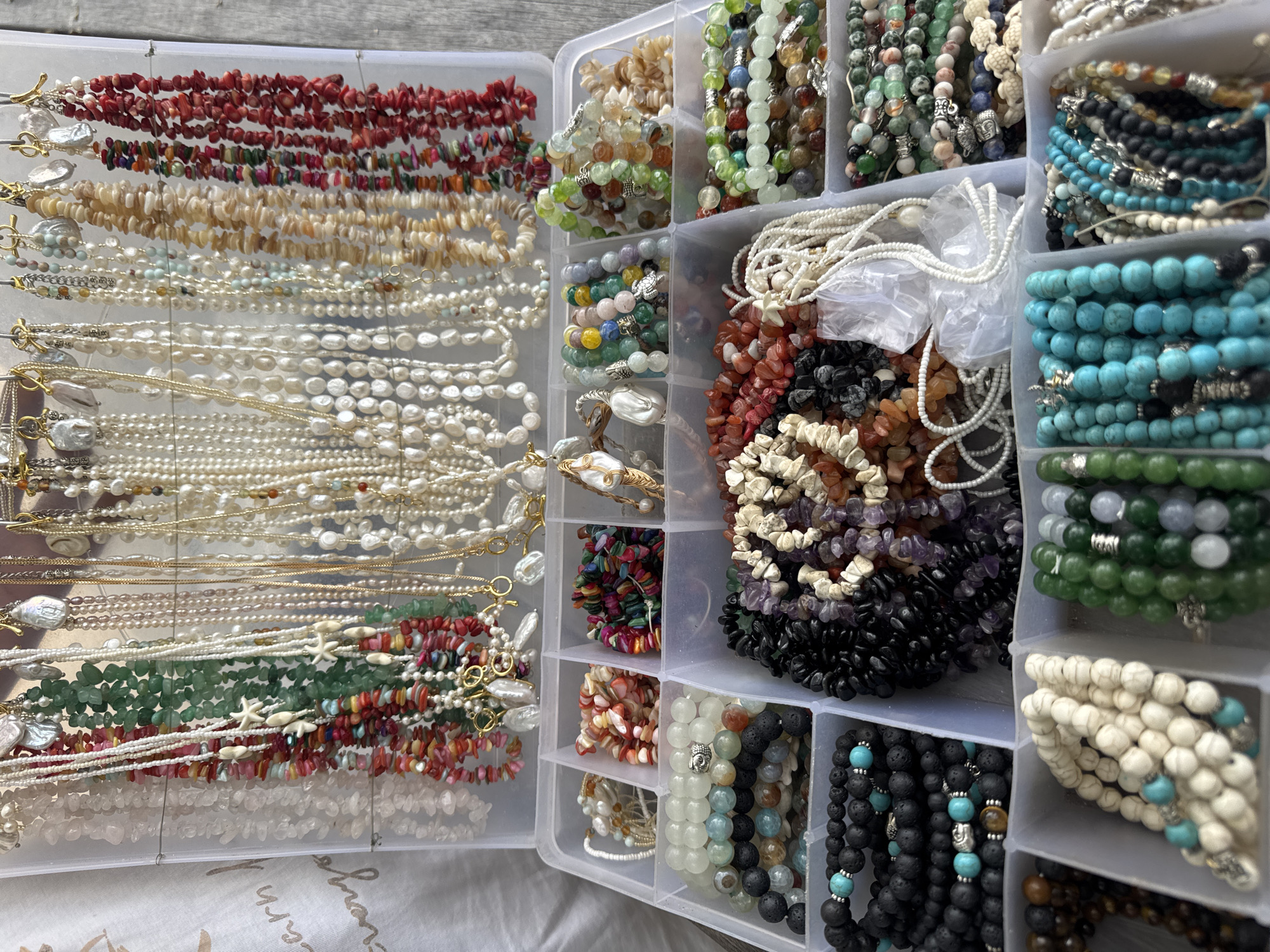 The image shows a collection of necklaces and bracelets organized in a clear plastic storage container with dividers. The jewelry appears to be made of various materials including beads, stones, pearls, and metal. Some of the necklaces have toggle clasps. The collection displays a wide range of colors and textures. The container is sitting on a white surface. The location is Unknown, Indonesia.