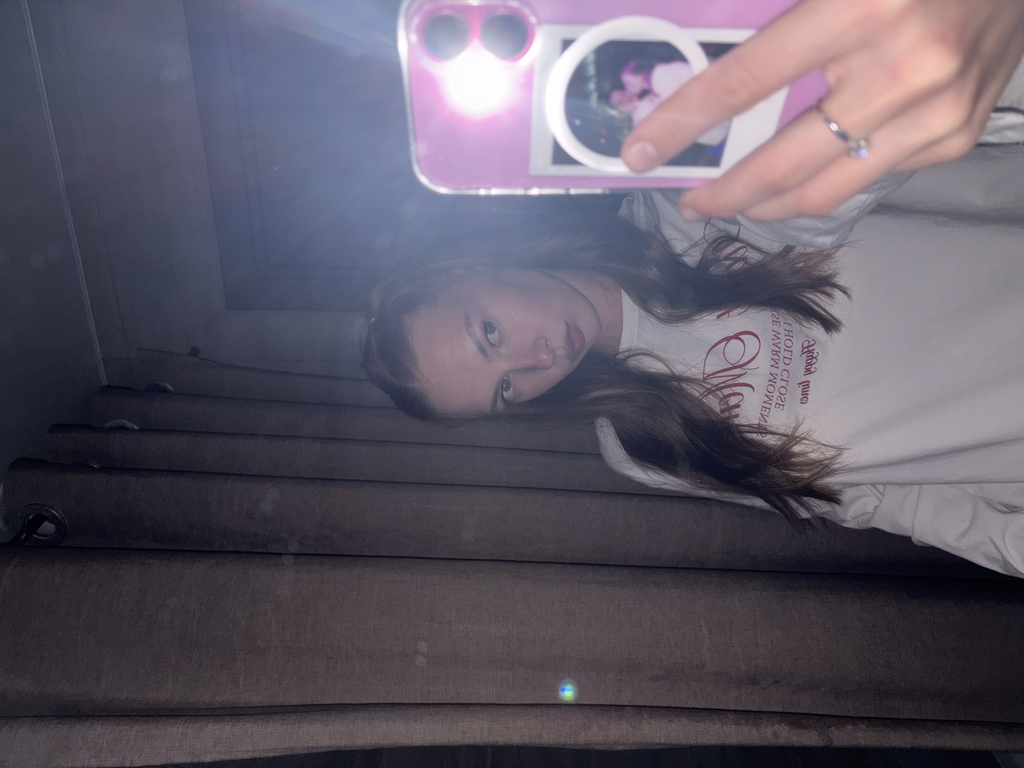 A young woman with her hair in a ponytail takes a selfie in a mirror. She is wearing a white long-sleeved shirt with red text on it. The text is reversed, indicating it is seen in a mirror. She is wearing a ring on her right hand, which is holding up a pink phone. The phone has a photo of a couple on its case. The background appears to be a bedroom with brown curtains. The scene is dimly lit, and there is a bright light glare from the phone. The image is likely taken indoors in Perm, Russia.