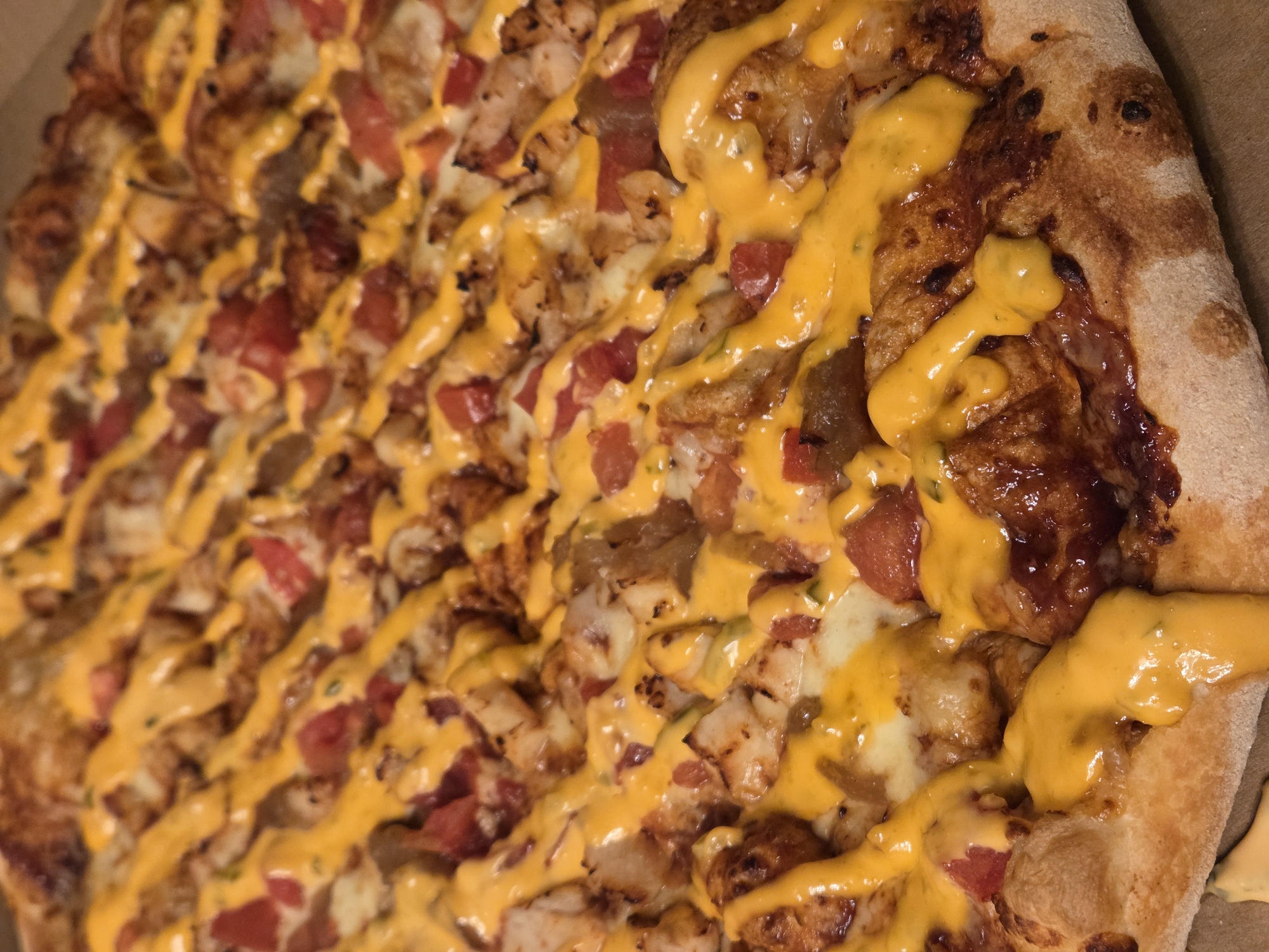 The image shows a close-up of a pizza in a cardboard box. The pizza has a thick crust and is topped with cheese, diced tomatoes, and pieces of what appears to be grilled chicken or meat. A yellow sauce, possibly a cheese sauce or a spicy mayonnaise, is drizzled across the top of the pizza. There are no people visible in the image. The location is unknown, and there is no indication that it is in Varna, Bulgaria.