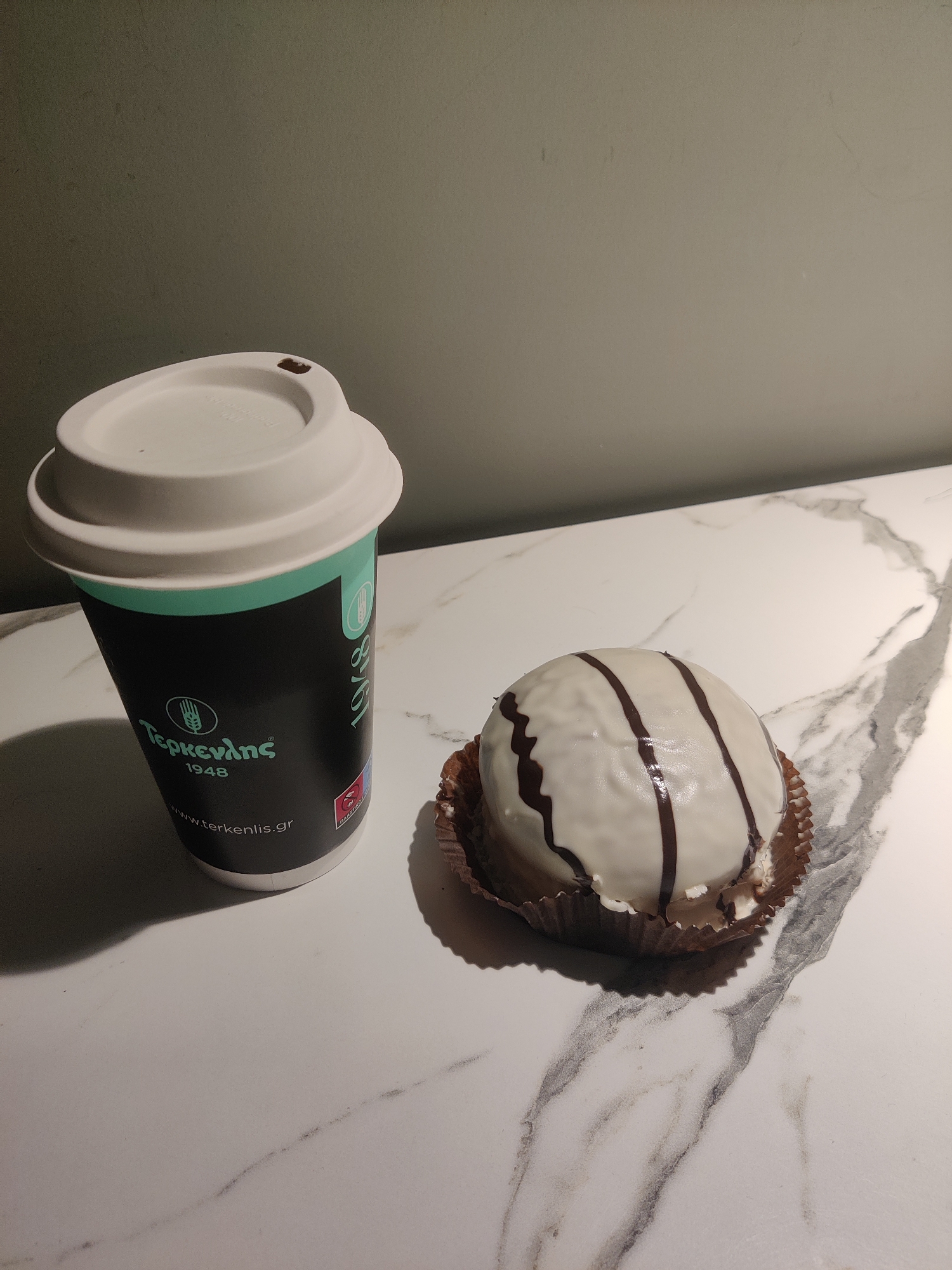 A white coffee cup with a teal and black design, featuring the text "Τερκενλης 1948" and "www.terkenlis.gr," sits on a marble countertop. Next to the cup is a round pastry, white-frosted with dark chocolate stripes, resting in a brown paper liner. The background is a muted gray wall. The scene appears to be in a cafe in Thermi, Greece, a suburb of Thessaloniki known for its high quality bakeries and cafes.