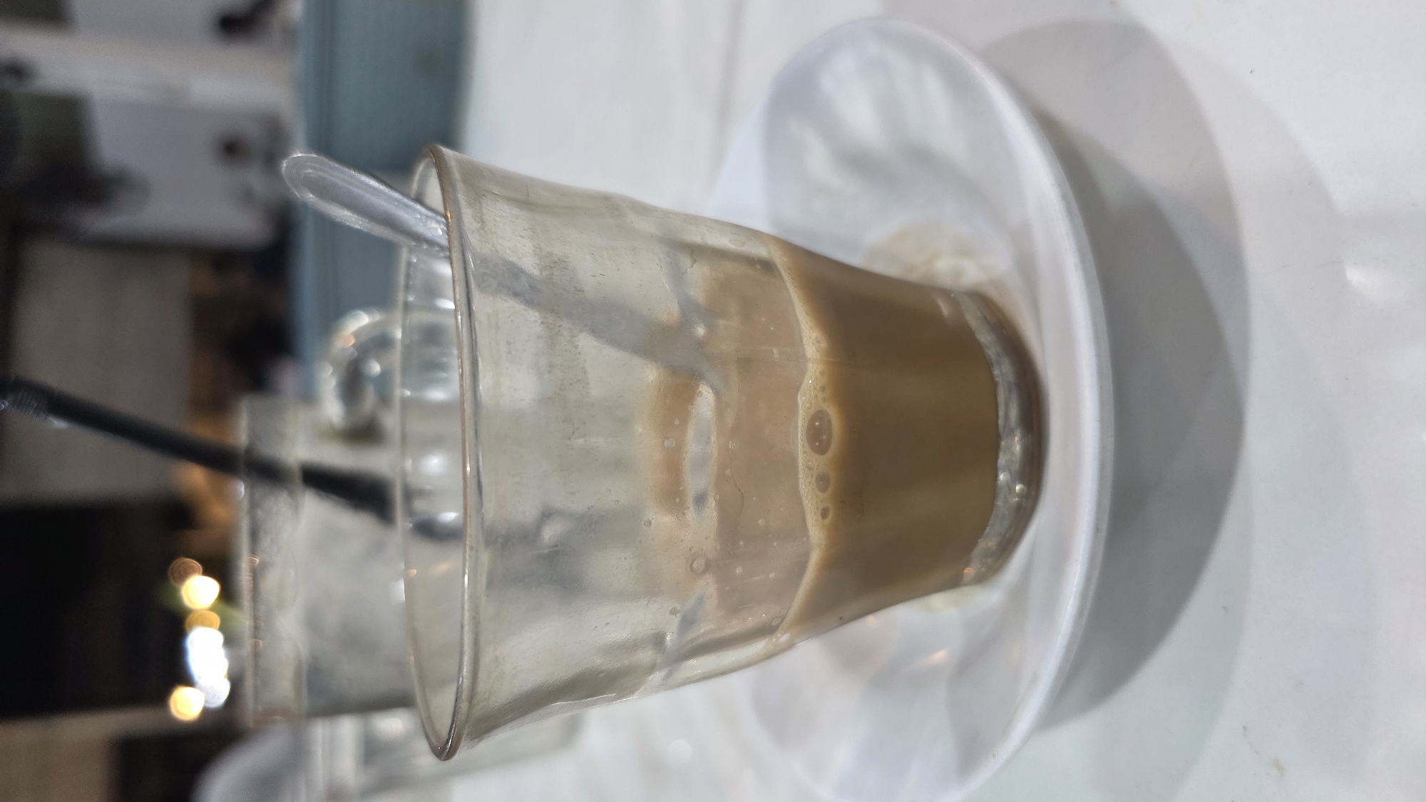 The image shows a glass of coffee with milk, half-empty, sitting on a white saucer. A metal spoon rests inside the glass. A black straw is visible in the background near another glass. The setting appears to be indoors, possibly a cafe or restaurant. The location is Lhokseumawe, Indonesia. The table has a white, slightly reflective surface. The background is blurred, but suggests an interior space with seating and lighting.