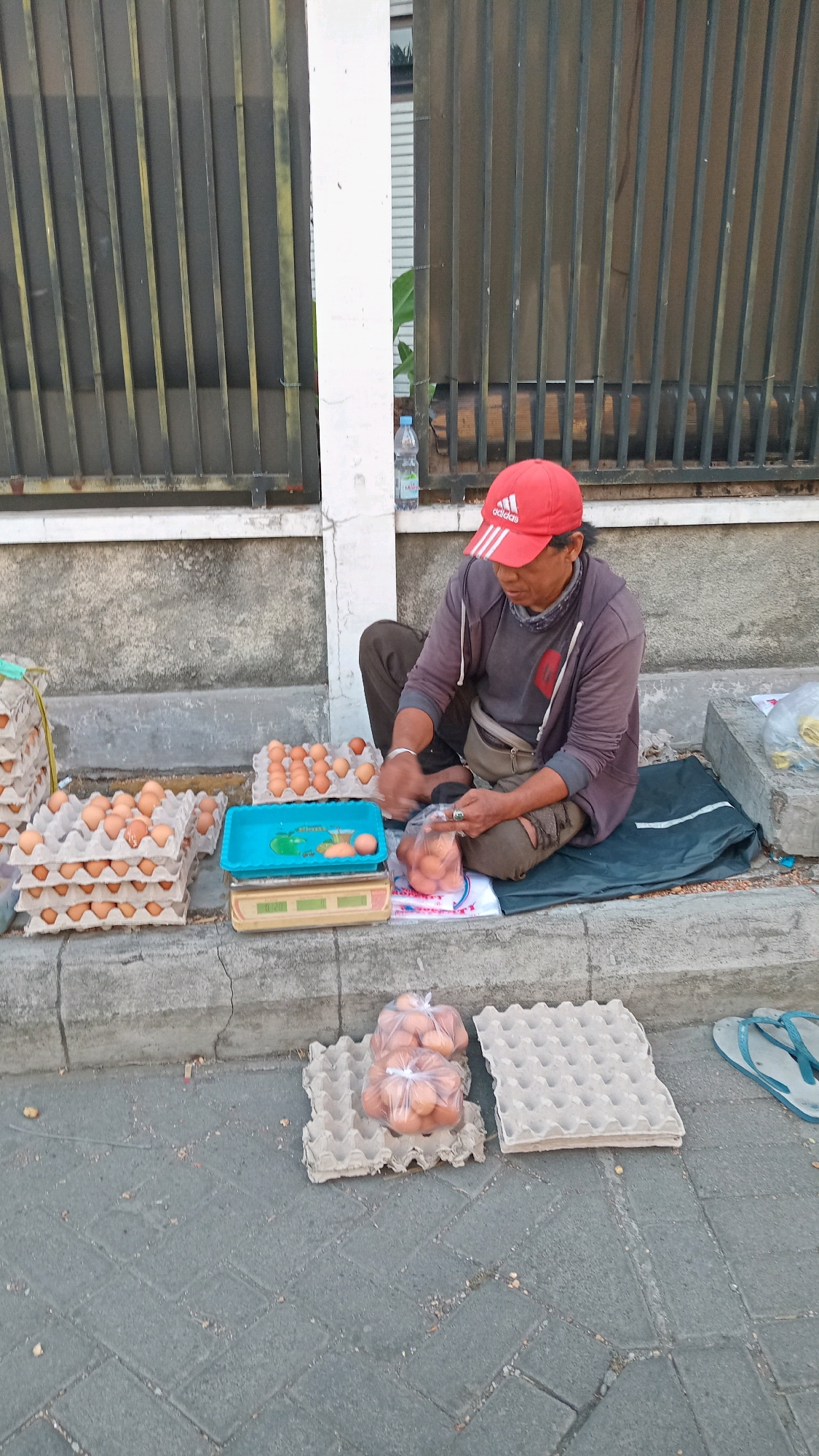A street vendor sits on the ground in front of a metal fence, selling eggs in the city of Randuagung, Indonesia. The vendor, wearing a red Adidas cap, grey hoodie, and dark pants, is surrounded by numerous cartons of eggs and plastic bags filled with eggs. A digital scale and a blue tray with some eggs are placed on the curb in front of the vendor. The ground is paved with grey interlocking stones. The scene appears to be set up for a street-side sale.