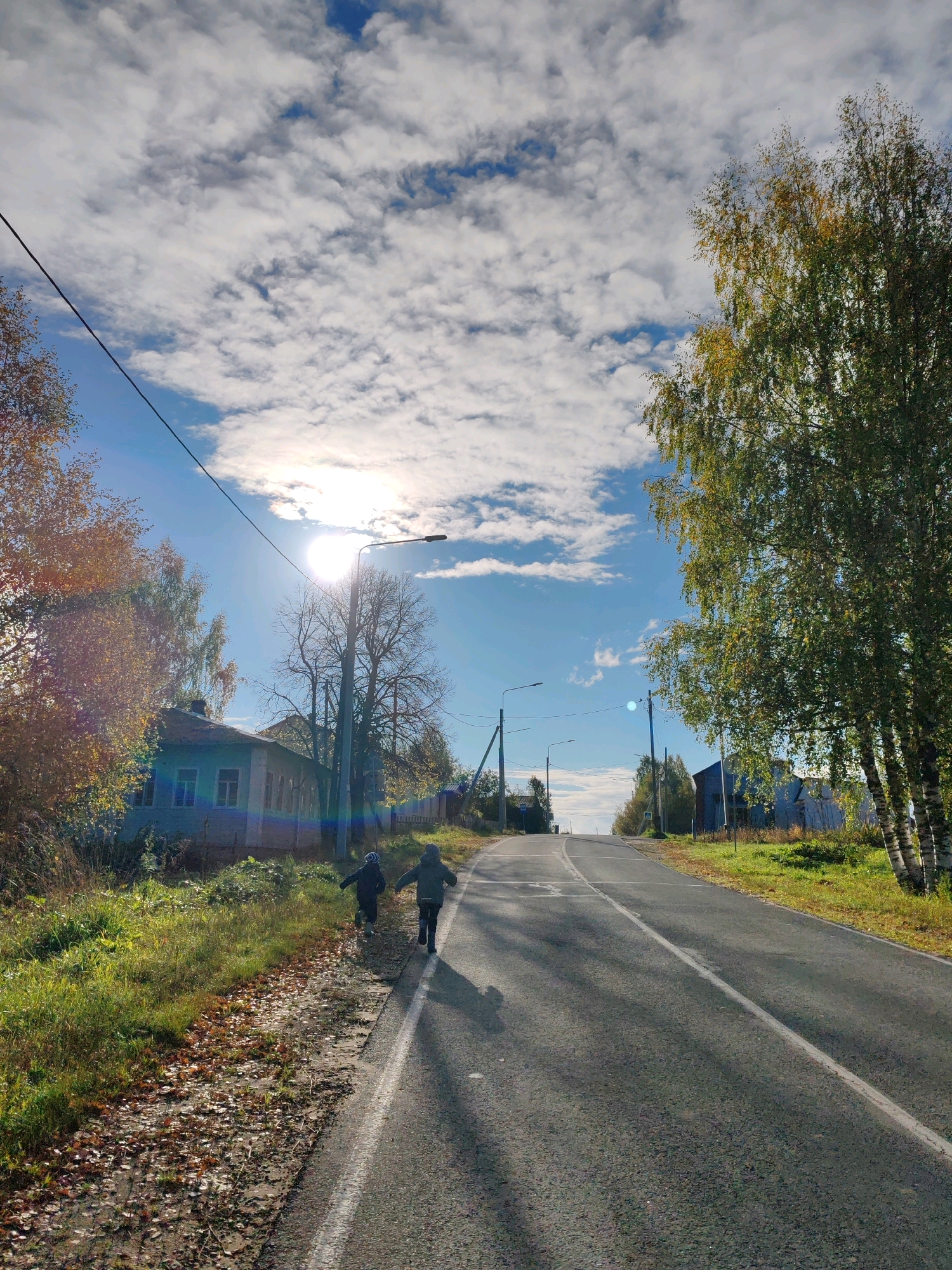 The image captures a rural road in Chukhlem, Russia, bathed in bright sunlight with scattered white clouds in a blue sky. The road, paved and with a white line marking its center, curves gently uphill and to the right. On the left side of the road, there is a grassy verge with fallen leaves and a small, light-colored house with several windows. A power line runs diagonally across the upper left portion of the frame. Several trees with green and yellowing leaves are present, particularly a cluster of birch trees on the right side of the road, their white bark visible. Two children, wearing dark clothing, are walking away from the camera on the left side of the road. The scene is illuminated by the sun, which is partially obscured by a cloud, creating lens flare. Streetlights are visible along the road, spaced at intervals. The overall impression is of a serene, autumn day in a village.