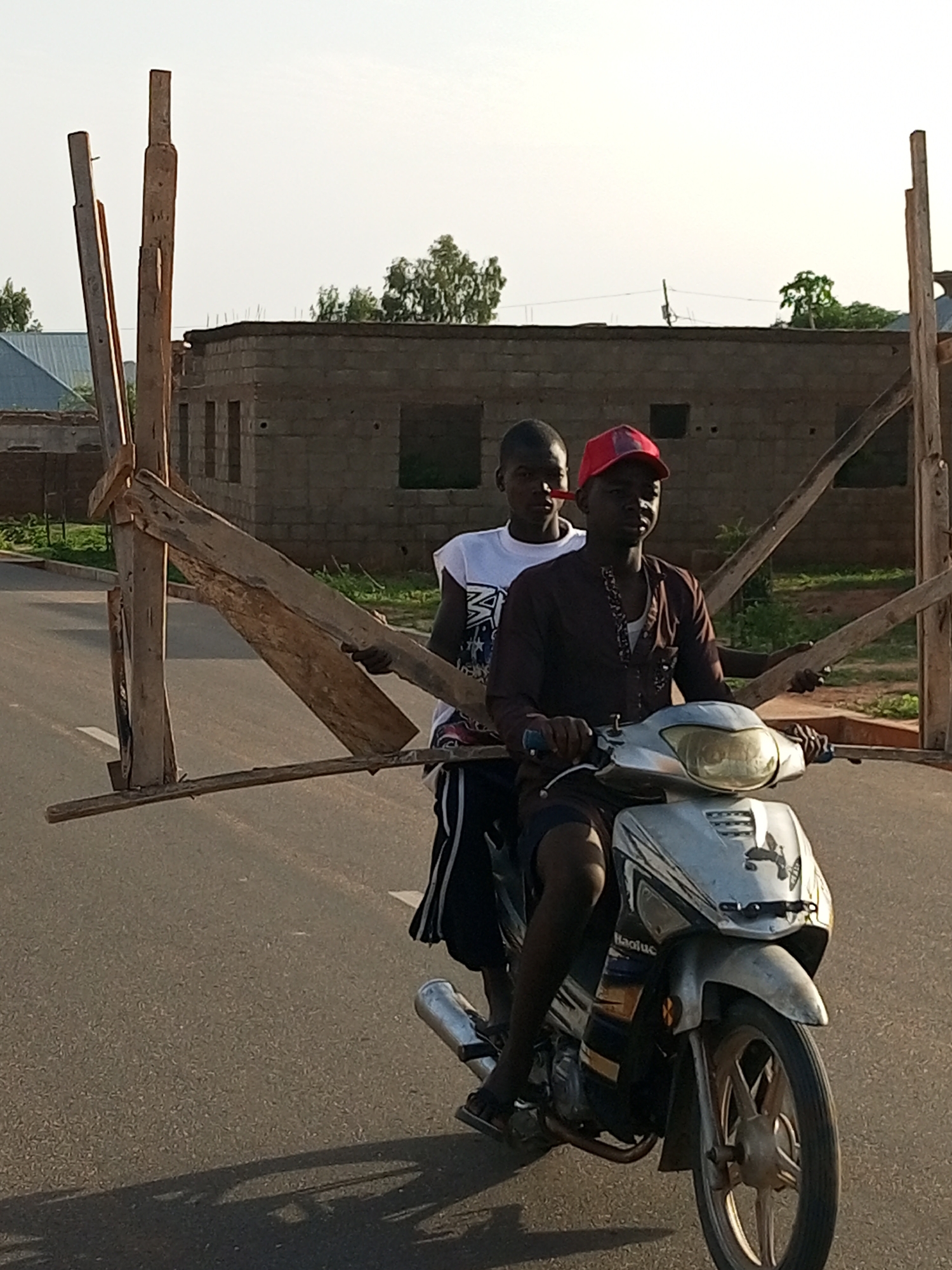 Two young men are riding a silver motorcycle on a paved road in Tempure, Nigeria. The man in the front, wearing a maroon patterned shirt and a red baseball cap, is driving. The man behind him, wearing a white t-shirt with graphics and dark striped shorts, is seated behind the driver. Both men are carrying a large, makeshift wooden structure that extends horizontally from the sides of the motorcycle, creating a wide load. The wooden structure appears to be made of rough-hewn planks and beams, held together by stakes and possibly ropes. In the background, there is a partially constructed building made of concrete blocks, with openings for windows and doors. Trees and vegetation are visible behind the building, and the sky is clear and bright. The road is asphalt with white lane markings. The scene suggests the men are transporting building materials or a dismantled structure.