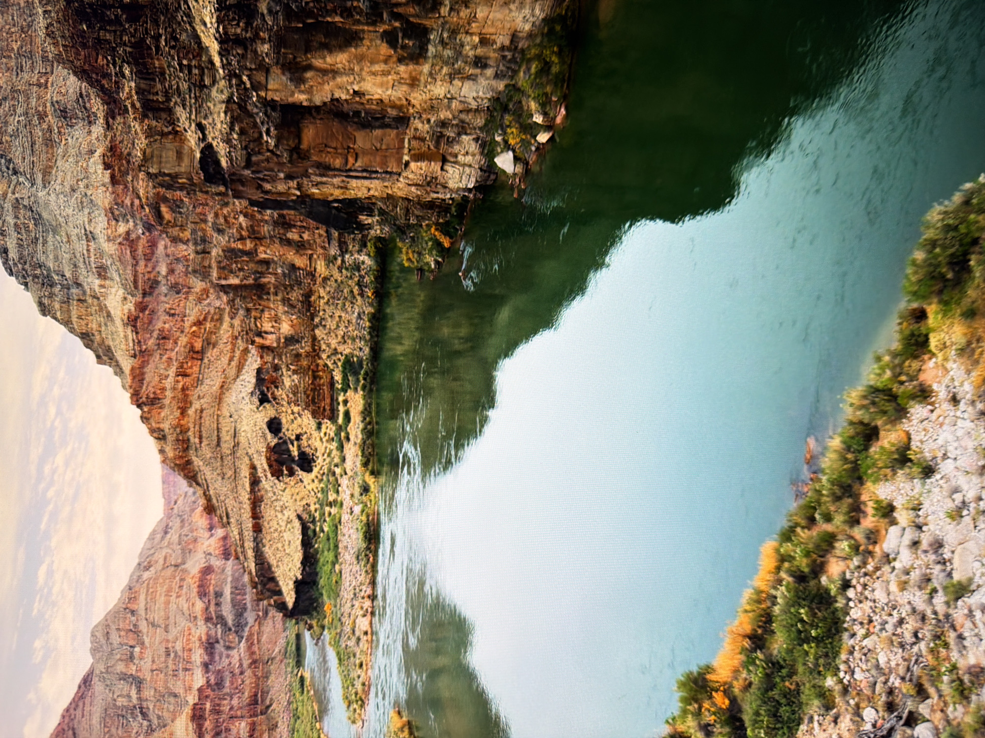 The image shows a wide river flowing through a canyon. The canyon walls are steep and made of layered rock, with a mixture of red, orange, and brown hues. Vegetation, including shrubs and grasses, grows along the riverbanks and on some of the lower slopes of the canyon. The river itself is a deep green color, reflecting the surrounding cliffs and sky. The sky above is partly cloudy with a soft, diffused light, suggesting it may be early morning or late afternoon. There are no discernible structures or human activity visible in the image, and it does not depict the city of Kyiv, Ukraine.
