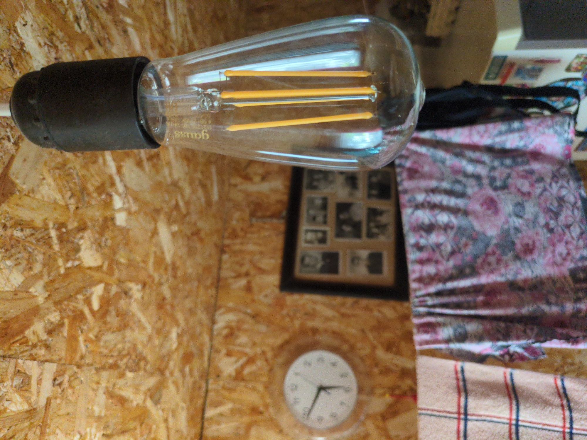 The image depicts an indoor scene, likely a shed or workshop, with walls constructed from oriented strand board (OSB). In the foreground, a clear Edison-style light bulb hangs from a black fixture. The bulb contains multiple yellow LED filaments. Behind the bulb, several items are visible. A round analog clock with a white face and black hands is mounted on the OSB wall, indicating approximately 9:15. To the right of the clock is a framed picture containing multiple black and white photographs of people. In the lower half of the image, two pieces of fabric are draped, one with a red and blue plaid pattern and another with a floral pattern in shades of pink and grey. The background suggests further clutter and unfinished construction. The specific location in Kulebaki, Russia, is not discernible from the visual evidence presented in the image.
