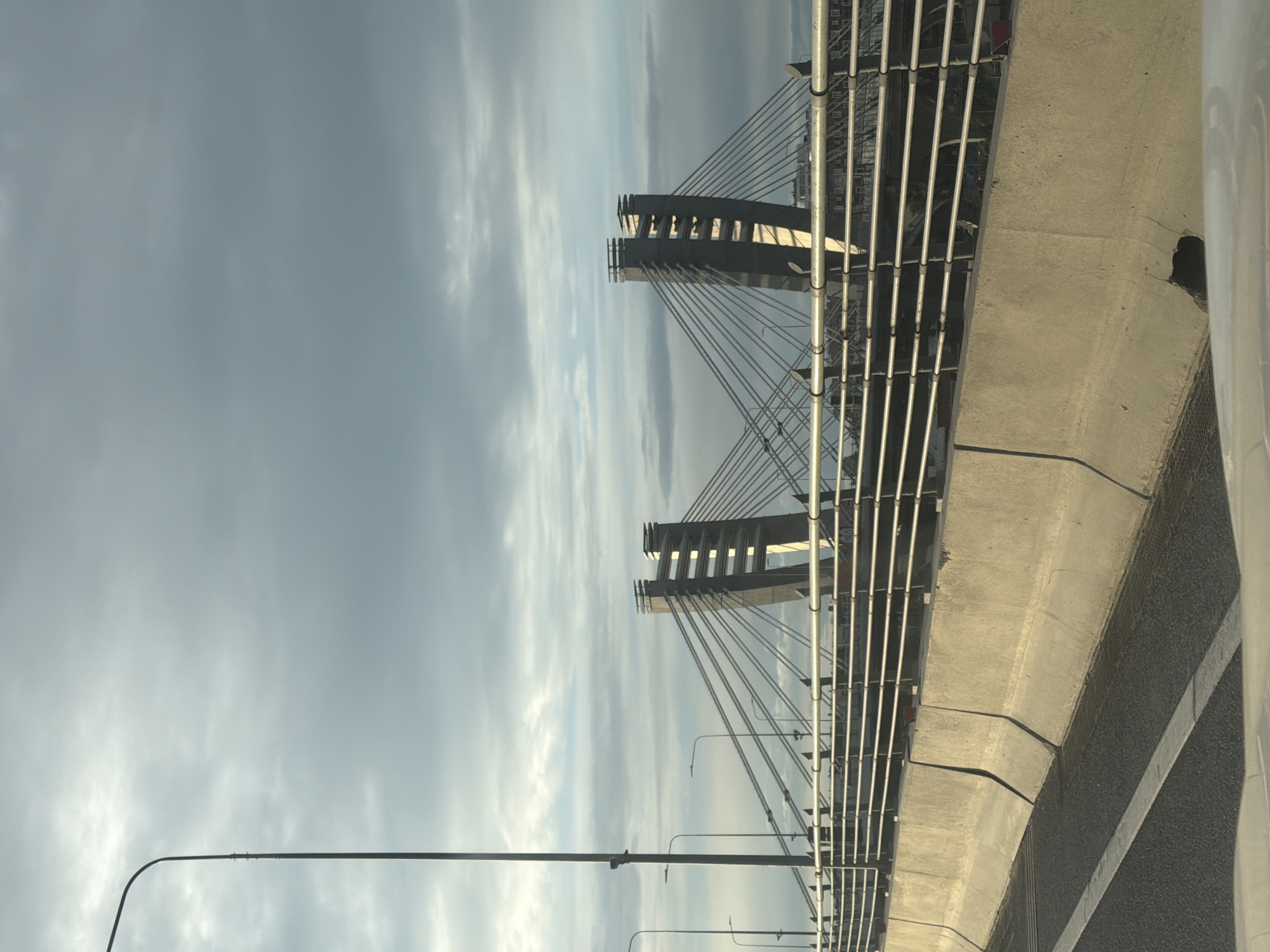 This is a photograph of a suspension bridge taken from a moving vehicle. The image is oriented vertically, with the sky occupying the upper left portion of the frame and the bridge structure dominating the right and lower portions. The sky is overcast with soft, diffused light, suggesting it is either early morning or late afternoon. A tall, slender lamp post curves into the frame from the bottom left, its dark silhouette contrasting with the lighter sky.
The bridge features two prominent, angled towers from which numerous cables descend to support the roadway. The towers are dark gray and appear to be made of concrete or a similar material. The cables are thin and numerous, creating a striking geometric pattern against the sky. A metal railing with vertical supports runs along the edge of the road, visible in the right half of the image. The road surface is visible in the bottom right, with a white line marking the lane. A concrete barrier runs along the side of the road, showing some wear and tear with a dark, shadowed opening visible in one section.
The overall mood of the image is somewhat stark and architectural, with a focus on the structural elements of the bridge and the vastness of the sky. There is no visible text in the image. The activity taking place is simply the passage over a bridge, as implied by the perspective from a moving vehicle.