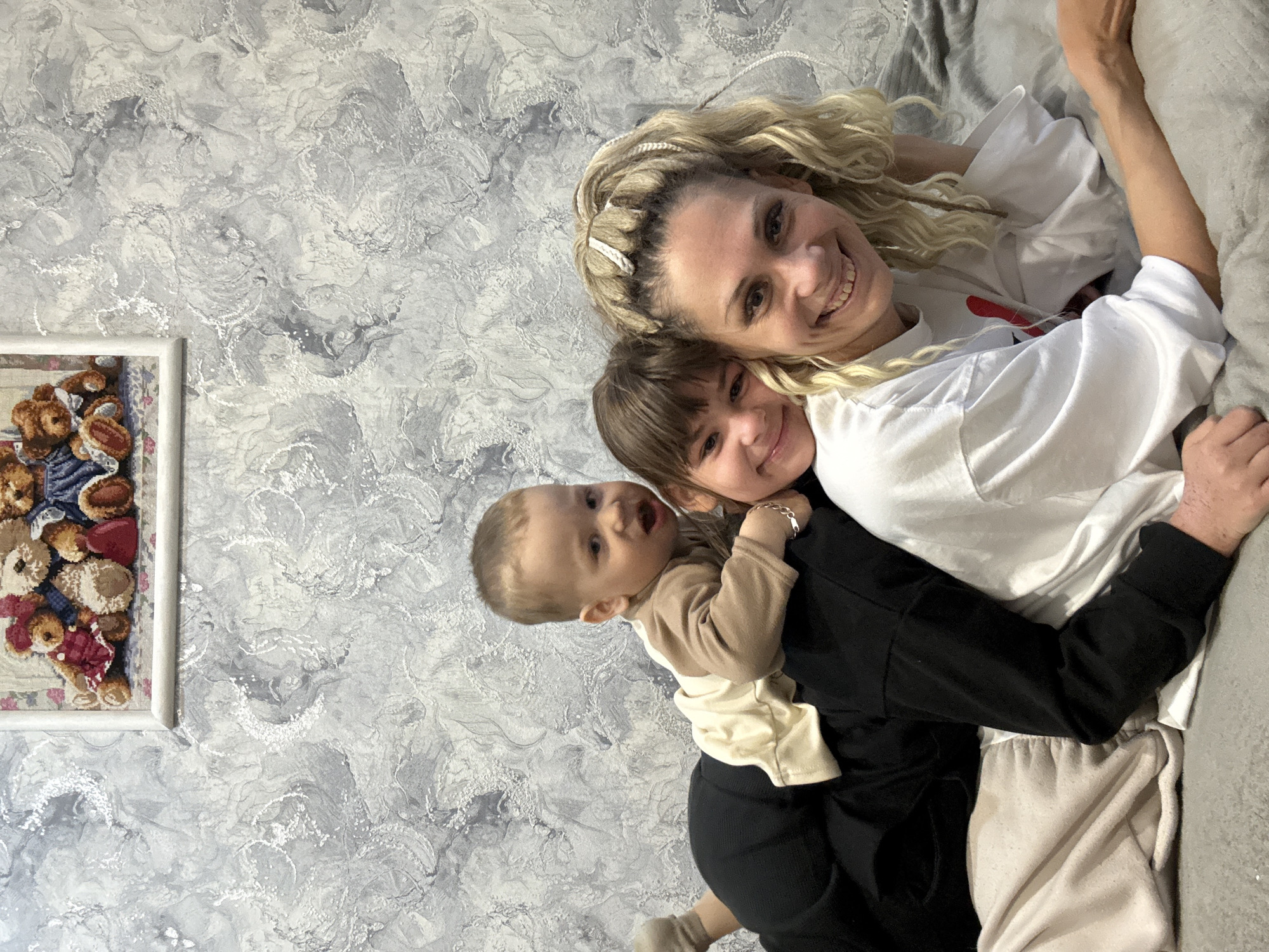 A woman with blonde curly hair smiles, lying on a gray couch and wearing a white t-shirt. A young girl with brown hair smiles while embracing the woman. A baby wearing a tan shirt is being held on the shoulders of a person wearing a black jacket. Behind them is a wall with a gray and white swirled pattern. A framed picture hangs on the wall, depicting teddy bears. The location is Rusanovo, Russia.