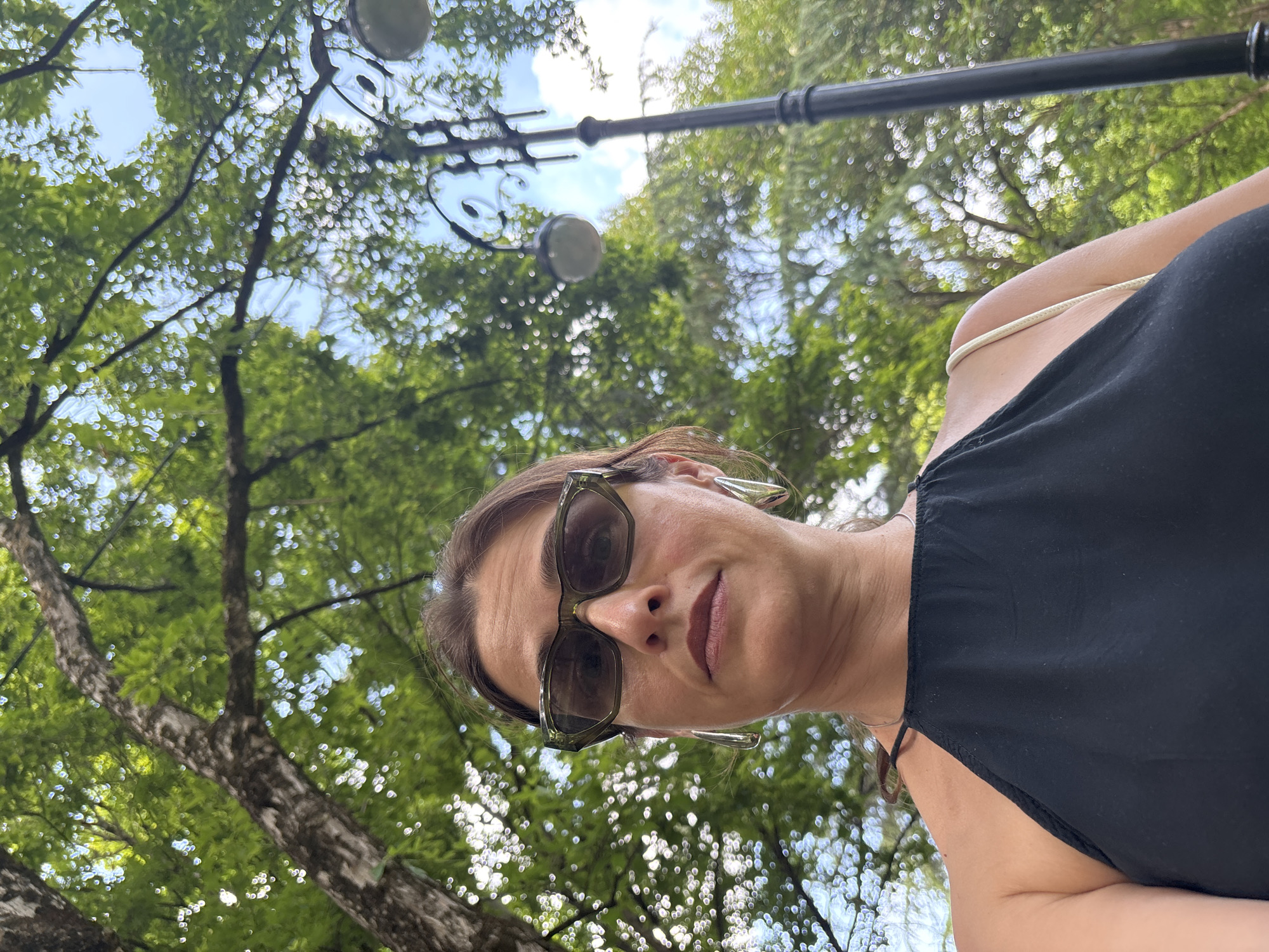 A woman is posing for a selfie outdoors. She is wearing a black halter top and sunglasses. The background is out of focus and shows trees and a lamppost, suggesting a park or public garden setting. The sunlight filtering through the leaves creates a bokeh effect. The image is taken from a low angle, looking up at the woman and the surrounding foliage.