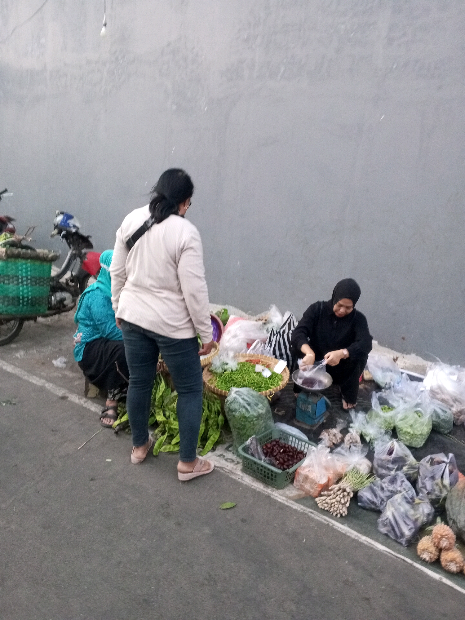 The image depicts an outdoor market scene on a paved surface, likely a street or roadside, in Gentan, Indonesia. A gray wall forms the background, with a single lightbulb hanging from a wire. In the foreground, various fresh produce is displayed.
On the right side of the image, a woman wearing a black hijab and dark clothing is kneeling and using a small blue scale. She is placing items into a metal bowl on the scale, appearing to weigh them. Around her are several plastic bags filled with produce, as well as baskets containing vegetables.
To the left, another woman is standing, facing away from the camera, wearing a light-colored jacket and dark jeans. She is partially obscuring a person sitting on a stool, who is wearing a bright turquoise headscarf and dark clothing. A motorcycle with a green basket attached is parked behind them.
The produce visible includes bunches of green beans, what appear to be chilies or small peppers in a green basket, and other vegetables and root crops in plastic bags and baskets. The overall impression is a typical street-side market where vendors sell fresh goods.