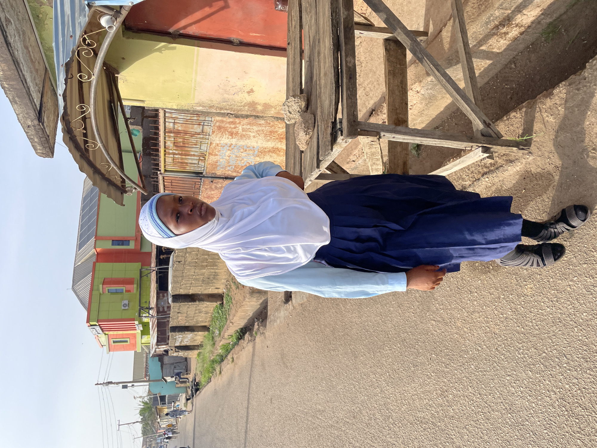 A full-body, horizontally oriented photograph depicts a young person standing on a paved surface, looking towards the camera with a slight smile. The individual, dressed in a white hijab, a light blue long-sleeved top, and a dark blue long skirt, appears to be of dark complexion. Dark, textured sandals complete the outfit.
To the person's right, a rustic wooden structure, possibly part of a stall, is visible, with a few light-colored stones resting on it. Directly behind the individual, a weathered building features a corrugated metal awning adorned with decorative metalwork, its walls a mix of faded yellow and reddish-brown. Further in the background and to the right, a more modern, multi-story building stands out with vibrant green and orange facades, and solar panels are visible on its roof. Air conditioning units are also mounted on its exterior.
The paved ground beneath the person is a light, earthy brown, extending into the distance where a street scene unfolds. Utility poles with overhead wires line the street, and faint outlines of other buildings and scattered vegetation can be seen. The sky is bright and clear, indicating a sunny day, likely late morning or early afternoon, as suggested by the shadows. The general atmosphere suggests a semi-urban or developing area in a warm climate. No discernible text is visible in the image or its crops.
