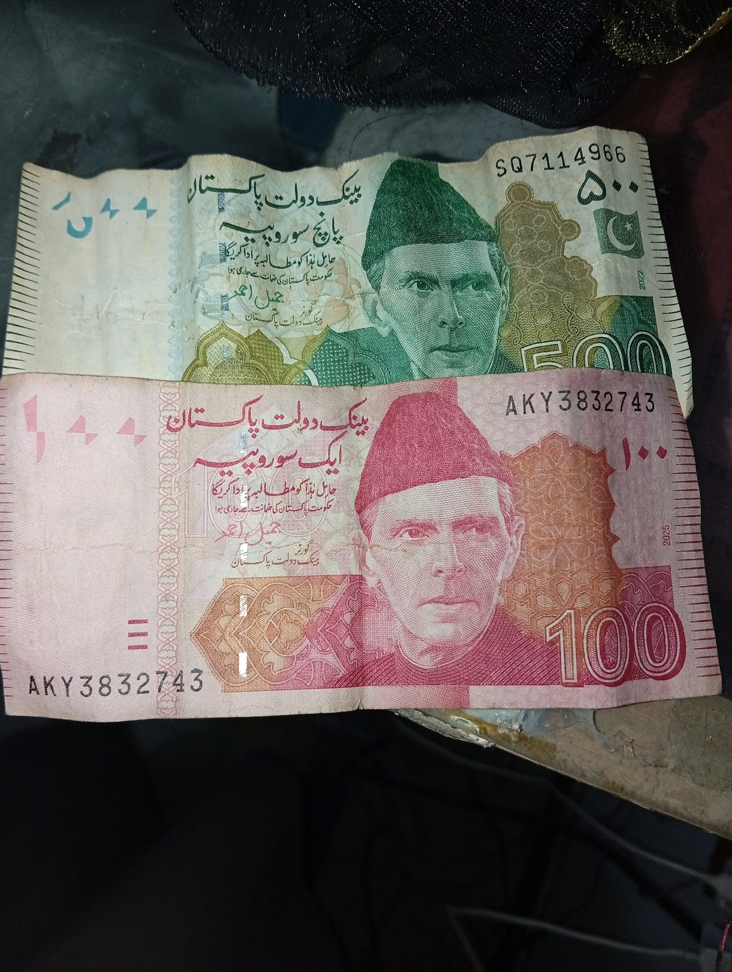 The image displays two Pakistani banknotes. The top banknote is green and features a portrait of Muhammad Ali Jinnah. Text on this banknote reads "500" and the serial number "SQ7114966". The bottom banknote is pink and also features a portrait of Muhammad Ali Jinnah. Text on this banknote reads "100" and the serial number "AKY3832743". Both banknotes have Arabic script printed on them, indicating they are legal tender in Pakistan. The banknotes are laid out on a dark, textured surface, possibly fabric or a tablecloth. There are no people or specific landmarks visible that would indicate a location in Larkana, Pakistan. The scene is a close-up of the banknotes.