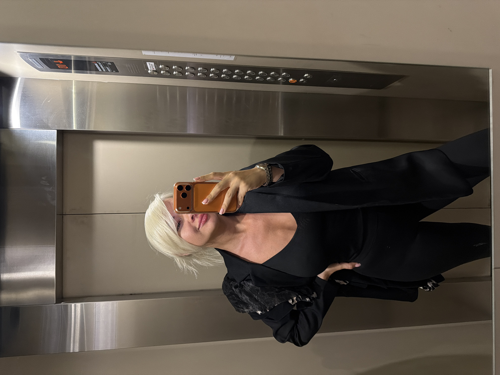 A woman with short blonde hair is taking a mirror selfie in an elevator. She is wearing a black V-neck top, a black blazer, and black leggings. The elevator has brushed metal walls and a panel of buttons on the right side. The elevator floor indicator shows "01" illuminated. She is holding an orange phone in her right hand and has her left hand on her hip.