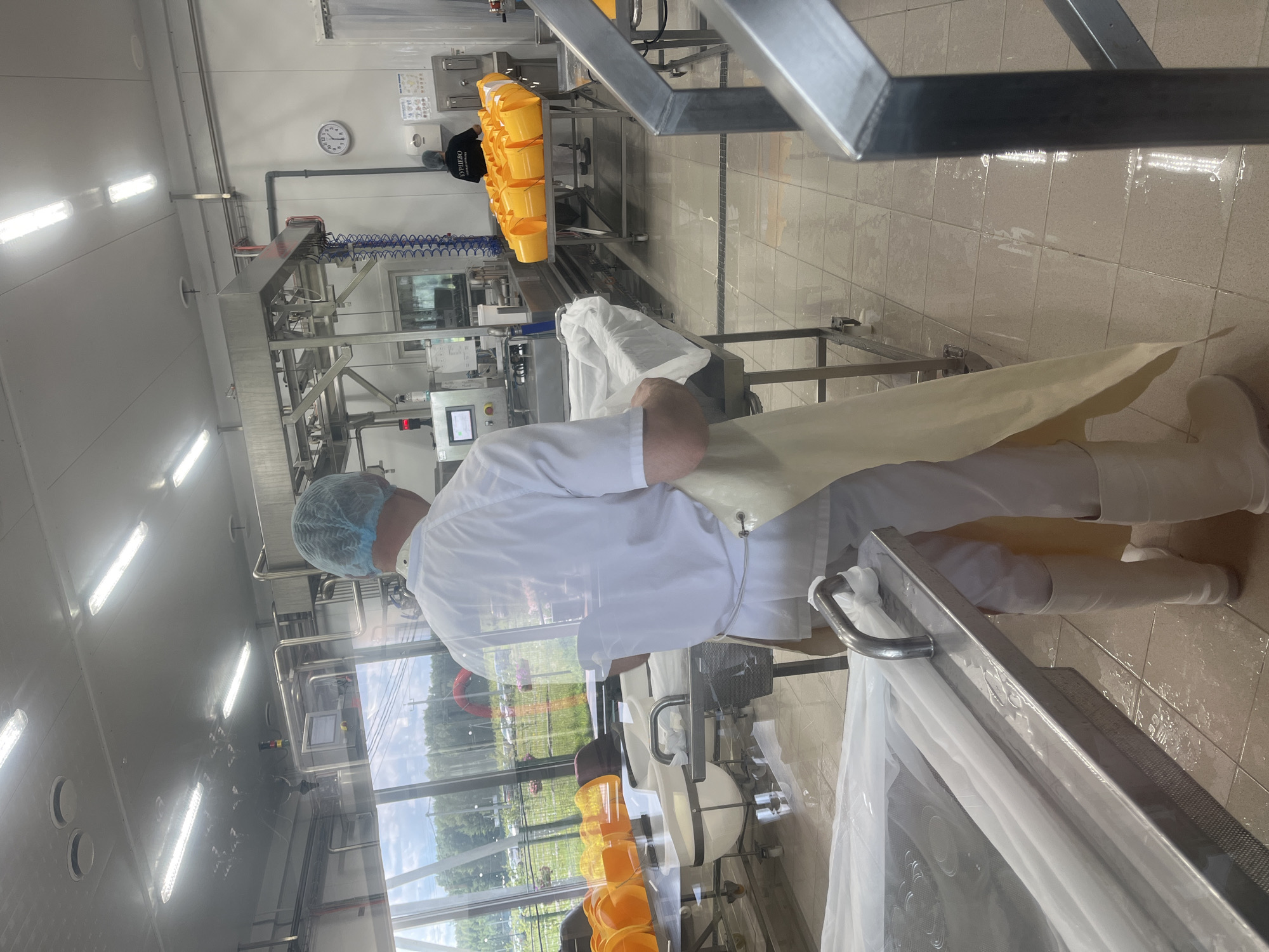 The image shows a man wearing a white uniform, a blue hairnet, and a yellow apron in a food processing facility. He is standing in front of a stainless steel table covered with white plastic sheeting, and appears to be working with it. The room has a tiled floor, industrial equipment, and large windows offering a view of a green outdoor area with trees and parked cars. Another person in a dark shirt with text is visible in the background, near a table laden with yellow buckets. A clock on the wall indicates it is approximately 11:44. The setting is consistent with a food production or preparation area in Akhlebaikha (Kovriginskii S/S), Russia.