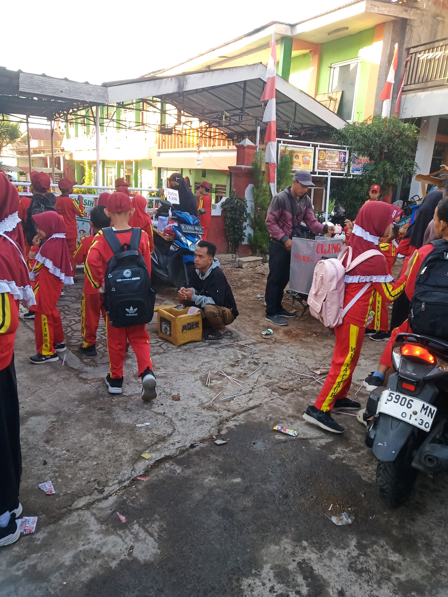 In Garut, Indonesia, a group of children, likely students based on their matching red and yellow uniforms with head coverings, are gathered outdoors. The scene appears to be in front of a school or community building, indicated by the presence of a multi-story building with green and orange accents in the background, and flags displayed. Several children are wearing backpacks, and some are interacting with an adult man operating a food cart selling "cilok." A motorcycle is parked nearby. The ground is paved with a rough, possibly asphalt or concrete surface, and there is some debris scattered around. The lighting suggests it is daytime, possibly late afternoon, with sunlight casting shadows.