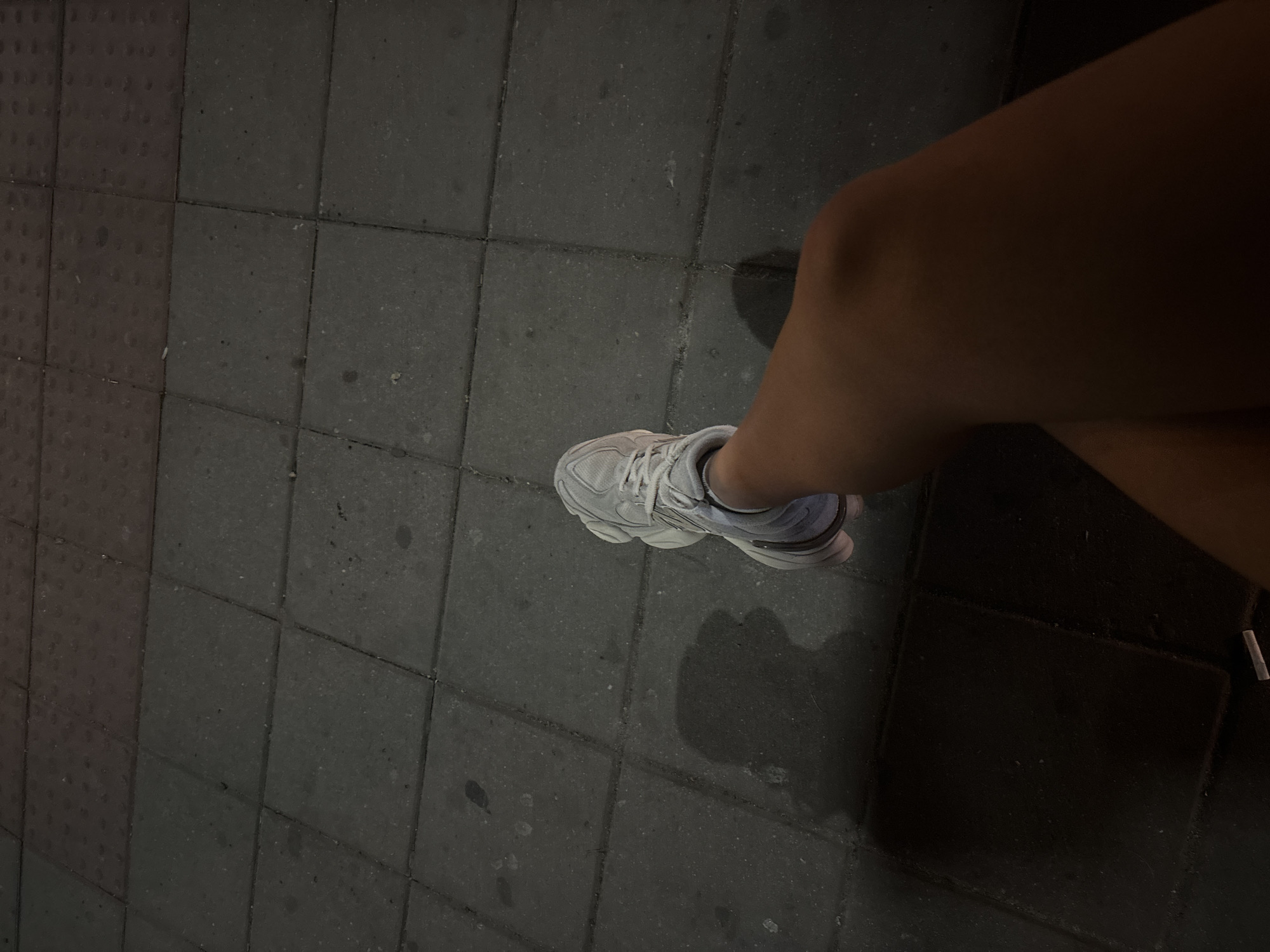 A low-angle, close-up, outdoor shot focuses on a person's lower leg and foot clad in a light-colored athletic shoe, resting on a gray tiled surface that appears to be a sidewalk or plaza. The lighting suggests it is nighttime or a dimly lit environment, casting a shadow of the person's leg and foot onto the tiles. The tiled surface exhibits subtle variations in color and texture, with some tiles appearing darker than others and showing minor imperfections. The background is dark and out of focus, hinting at the general urban surroundings. The image does not provide specific clues to pinpoint its location within Varna, Bulgaria, beyond being an outdoor urban setting with tiled ground.