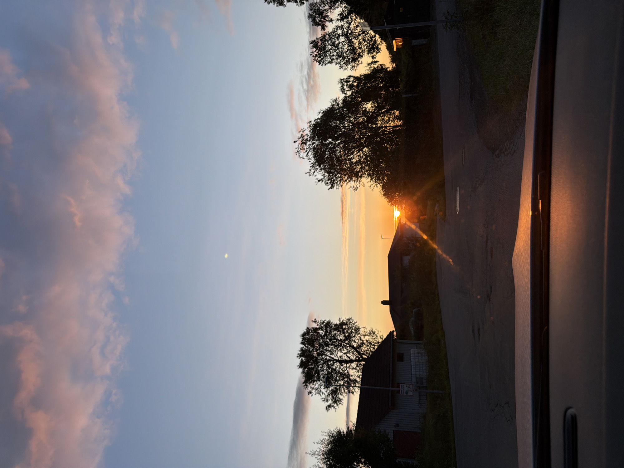 A frontal, low-angle, full shot captures a street scene in Harstad, Norway, at sunset. The sky is a gradient of pale blue transitioning to a soft orange glow near the horizon, with streaks of pinkish clouds scattered across the upper portion. A faint, small crescent moon is visible in the upper right quadrant of the sky.
In the middle ground, the setting sun, a bright orb, is partially obscured by trees and houses, casting a strong orange light and visible sun flares across the scene. The light creates lens flare effects on the pavement. Two houses are visible; one on the left is a light-colored, low-slung building with a dark roof, and another on the right has a darker roof and appears to be nestled behind trees. A tall, leafless tree stands silhouetted against the sky in the mid-left of the frame, near the first house. The road, which appears to be asphalt, curves gently from the left foreground towards the right middle ground, with some cracks and imperfections visible on its surface.
In the foreground, the top of a car's dashboard is visible, reflecting the warm sunset light. The dashboard is a dark, matte surface. The windshield is implied, with the view of the street and sky beyond it. The overall impression is a serene, natural landscape at dusk, likely a residential area on the outskirts of Harstad. There are no people visible in the image.