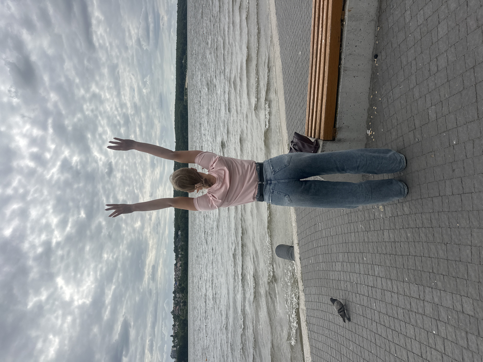 A full shot from behind shows a young woman with blonde hair, wearing a pink t-shirt and loose-fitting blue jeans, standing on a paved area near a body of water. Her arms are raised above her head, palms facing forward. The water is rippling, with a line of trees and buildings visible on the opposite shore, which is a park-like setting that is likely Ternopil's embankment. A pigeon stands on the paving stones to the left of the woman. A wooden bench is situated behind her, and a dark-colored bag rests on the ground beside it. The sky is overcast with clouds.