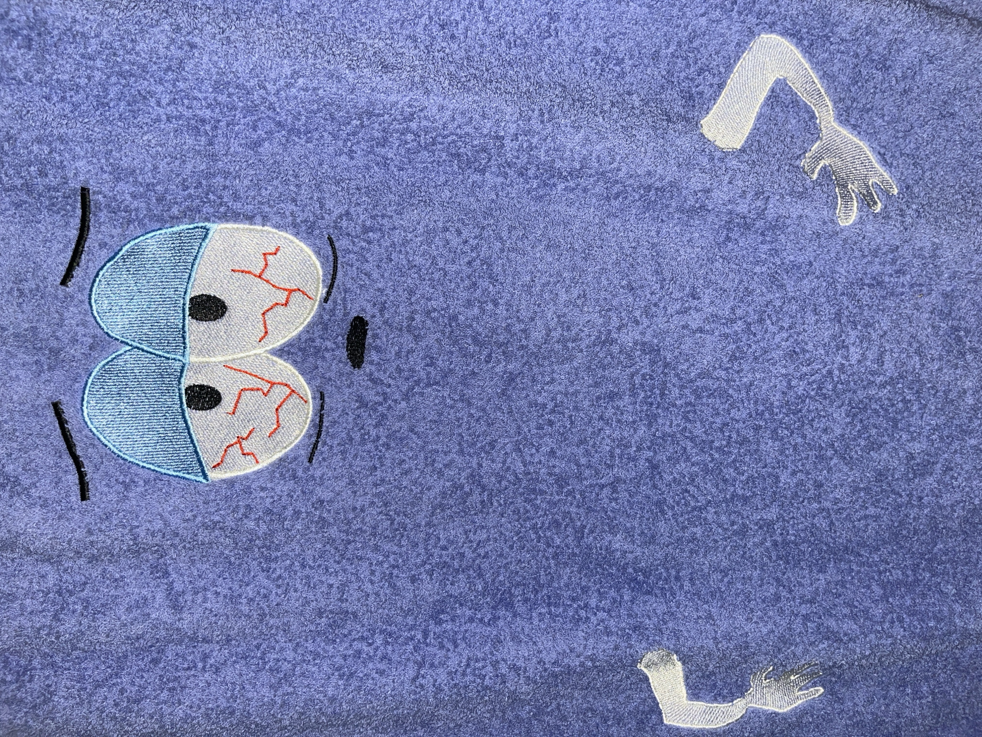 A close-up, high-angle shot shows an embroidered cartoon character on a purple, textured fabric, possibly a towel. The character has large, drooping eyes with black pupils and red-veined white scleras, suggesting tiredness or intoxication. Above the eyes are thin black lines representing eyebrows, and below is a small, downturned mouth. Two disembodied, embroidered arms extend from the sides of the frame. The left arm is bent and has a small, open hand with three fingers. The right arm is also bent, and its hand has four fingers. The fabric's texture is visible throughout the image, with a visible thread pattern from the embroidery. The scene is devoid of any discernible environmental context, rendering any attempt to pinpoint its location within a city impossible.