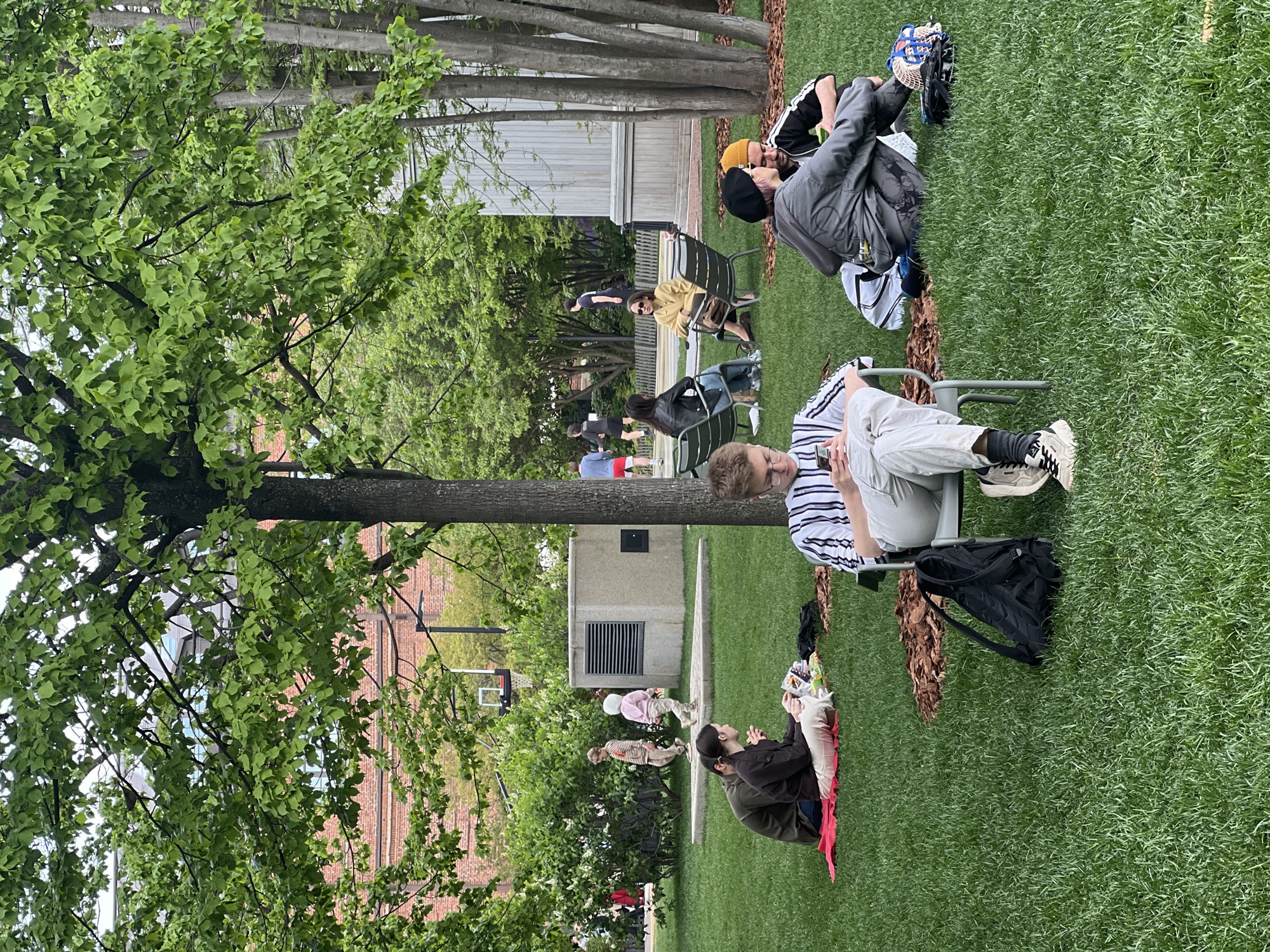 The image depicts an outdoor scene in a park-like setting. Several individuals are seated in chairs spread across a grassy area, seemingly relaxing or engaging in personal activities. Some are looking at their phones. There is a group of people sitting on a red blanket. Trees are present throughout the scene, and there are some buildings visible in the background. Based on the overall atmosphere and the presence of certain architectural elements, it may be in Saint Petersburg, Russia, though specific details such as landmarks are not distinctly visible to confirm the location.