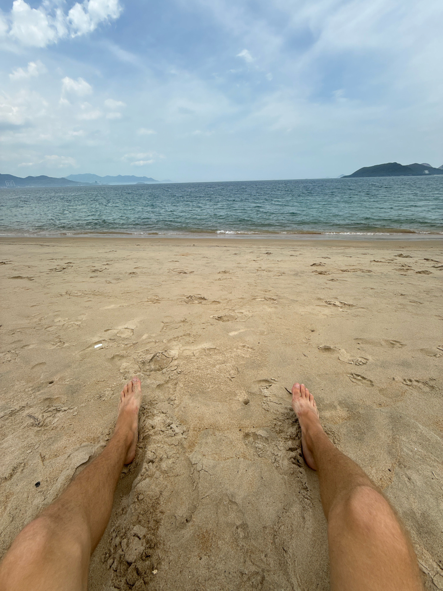 The image shows a person's legs and feet resting on a sandy beach, with the ocean and distant islands in the background. The sand is light brown with footprints scattered across it. The ocean water is a deep blue with gentle waves. The sky is partly cloudy, with white clouds against a pale blue backdrop. In the distance, several islands or peninsulas are visible, some with hazy mountain formations. The scene appears to be a tranquil beach setting in Nha Trang, Vietnam, possibly on a day with moderate cloud cover.