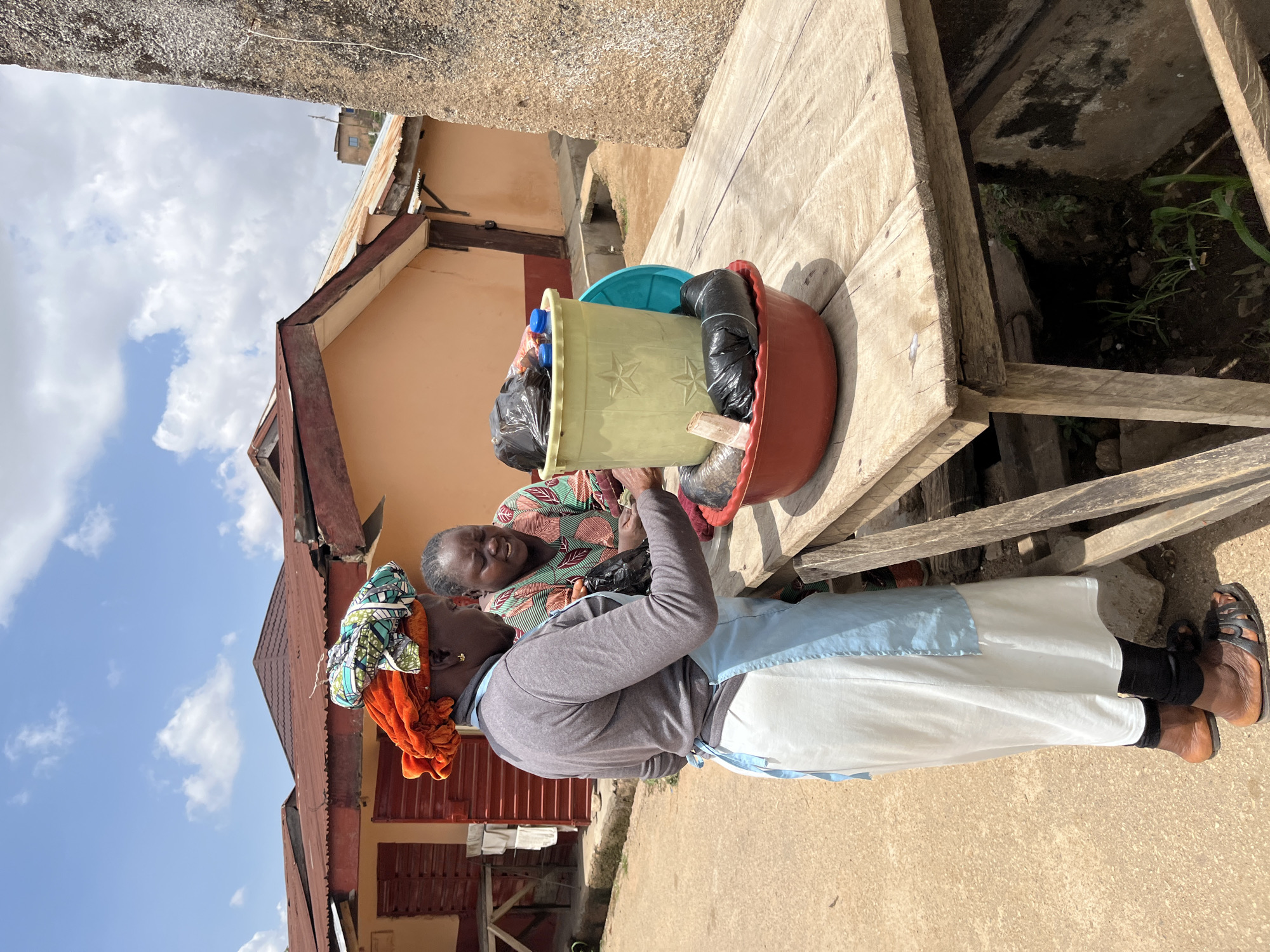 Two women are standing at a wooden table outdoors. The woman on the left is wearing a gray long-sleeved shirt, a white apron with light blue trim, a colorful headscarf, and sandals. She is leaning over the table, interacting with the woman on the right. The woman on the right is wearing a colorful patterned shirt and is also leaning towards the table. In front of them on the table are two plastic containers and a red bowl filled with what appear to be packaged items wrapped in black plastic. A bottle of water with a blue cap is visible in one of the containers. The background shows buildings with corrugated metal roofs and orange-colored walls, typical of the urban landscape in Jos, Nigeria. The ground is a dirt or gravel surface. A large, rough-textured stone structure is visible to the right of the frame, possibly part of a building or natural formation. The sky is partly cloudy.