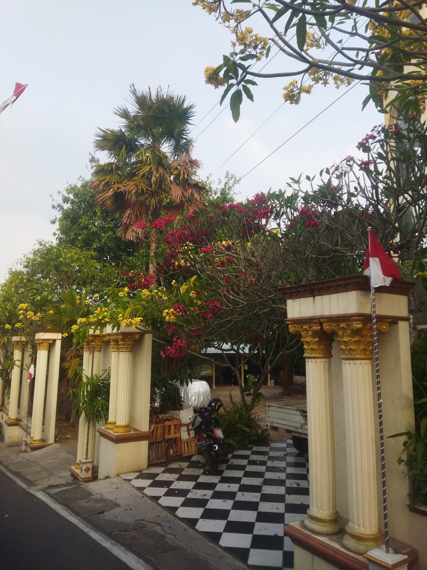 A motorcycle is parked on a black and white checkered tile floor next to a gated entrance with ornate pillars. The entrance is adorned with the Indonesian flag. Lush greenery, including palm trees and flowering plants with red and yellow blossoms, surrounds the entrance. The scene appears to be a residential or public area in Wonogiri, Indonesia, likely a street-facing property. The sky is overcast, suggesting a daytime setting.