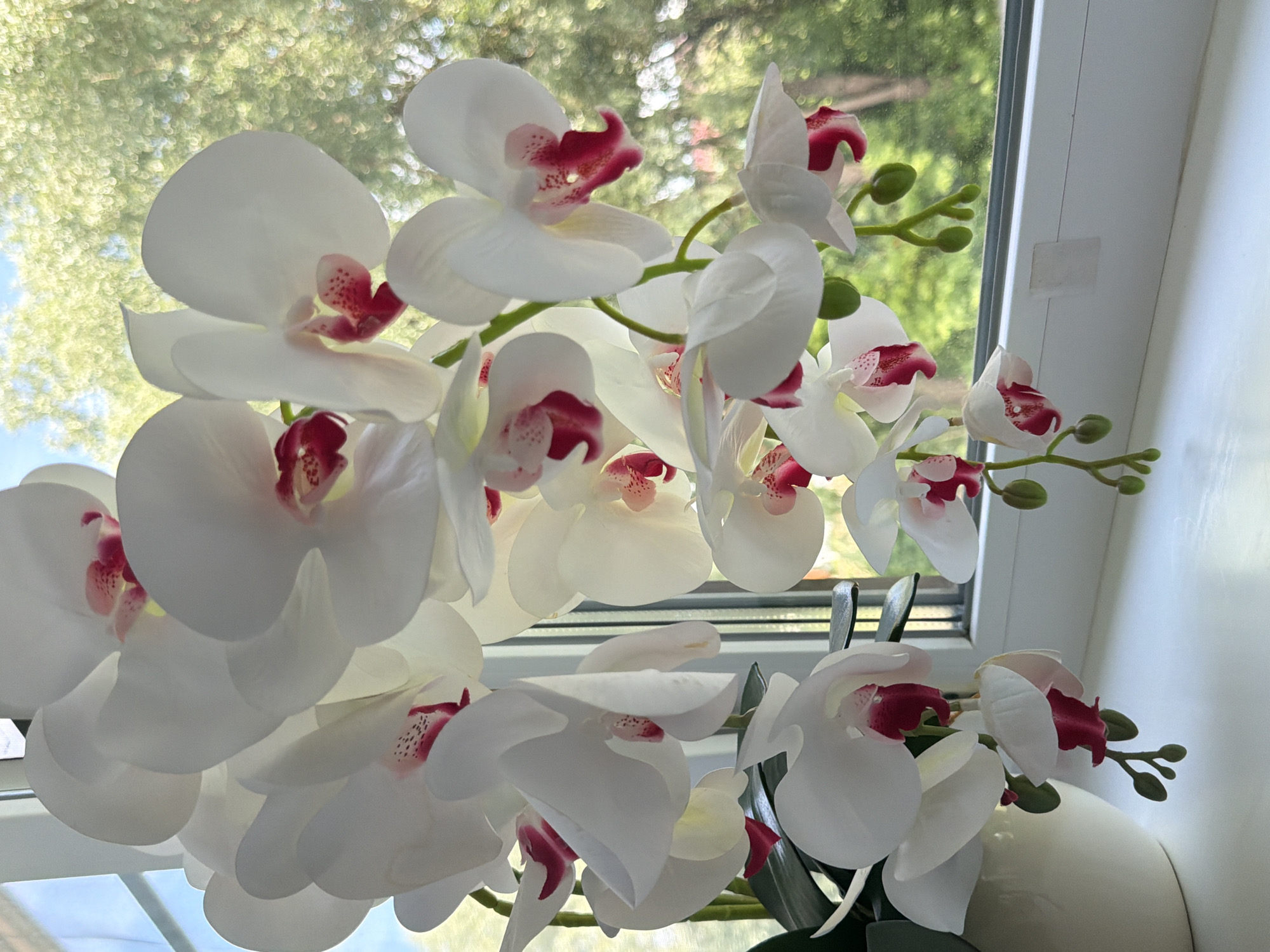 A close-up, eye-level shot captures a bouquet of artificial white orchids with deep pink centers and speckled accents, arranged in a light-colored ceramic pot. The orchids are positioned on a white windowsill, with the lush green foliage and bright sky of an outdoor environment visible through a window in the blurred background. The scene is brightly lit, suggesting daytime.