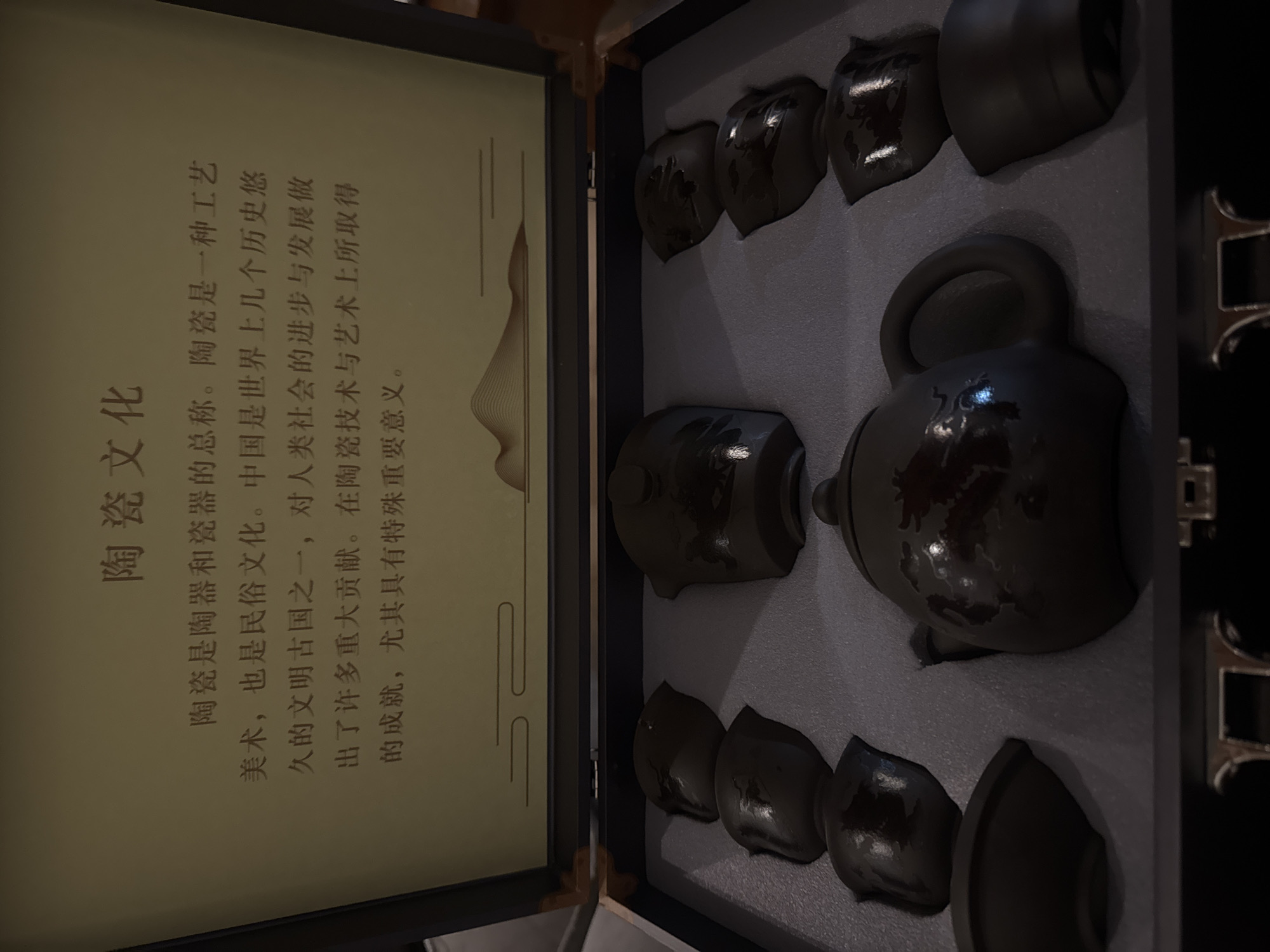 An overhead shot captures an open display box featuring a dark-colored ceramic tea set and a framed informational text. The tea set, neatly nestled in a grey foam insert, comprises a prominent teapot with a handle and spout, surrounded by eight distinctive teacups. The ceramic pieces are dark, possibly black or very dark grey, with subtle reflective highlights suggesting a glazed finish and intricate, abstract patterns. The teacups feature a unique, slightly angular design, wider at the top and tapering down.
On the left side of the box, a framed piece of text, written in Chinese characters on a light beige background with decorative wavy lines, explains the cultural significance of ceramics. The text is titled "陶瓷文化" (Ceramic Culture) and describes ceramics as a form of industrial art and folk culture. It highlights China's role as an ancient civilization that has made significant contributions to human progress through its ceramic technology and art, emphasizing the special importance of these achievements.
The scene suggests an indoor setting, likely a retail display, museum exhibit, or personal collection, showcasing items of cultural value. No human subjects or activities are present. The image was captured in Kuta Selatan, Indonesia, indicating the location of this cultural display.