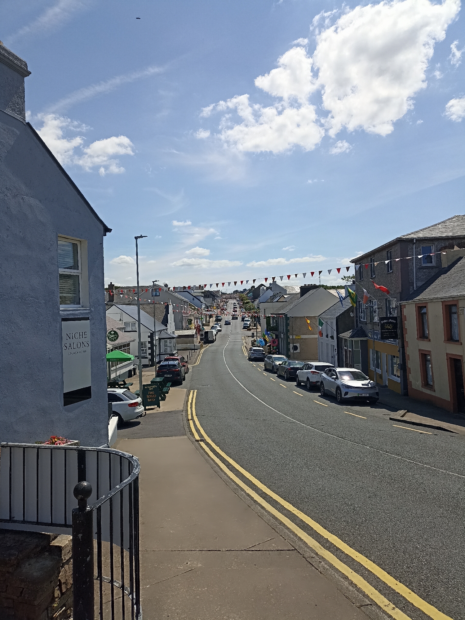 The image captures a street view in Dungloe, Ireland, likely on a sunny day with scattered clouds. The foreground is dominated by a grey building on the left, featuring a white sign for "NICHE SALONS." Adjacent to it is a black metal railing. The street, paved with asphalt and marked with double yellow lines, curves to the right, lined with various businesses and residential buildings. Many of these buildings display flags, including Irish flags, and red and white triangular bunting stretches across the street, suggesting a festive occasion. Several cars are parked along the street and on the opposite side of the road, with more vehicles visible in the distance, indicating moderate traffic. The architecture appears typical of a coastal Irish town.