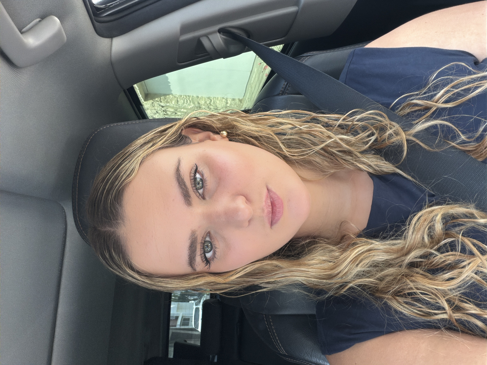 A young woman with blond, curly hair is in the driver's seat of a car, wearing a blue shirt and a seatbelt. She is looking directly at the camera. The interior of the car is visible, including the dashboard, windows, and headrest. The exterior appears to be a neighborhood. The location is undetermined in San Bernardino Tlaxcalancingo, Mexico.