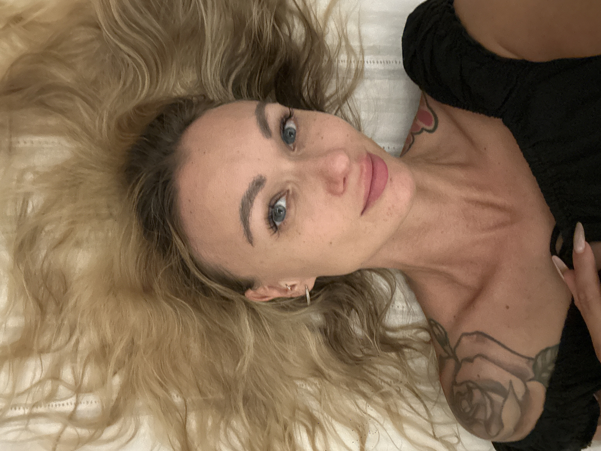 A woman with blonde hair and blue eyes is lying on a white surface. She is wearing a black top. Tattoos are visible on her shoulder and chest. She is looking directly at the camera. The location is unknown and not provided within the image.