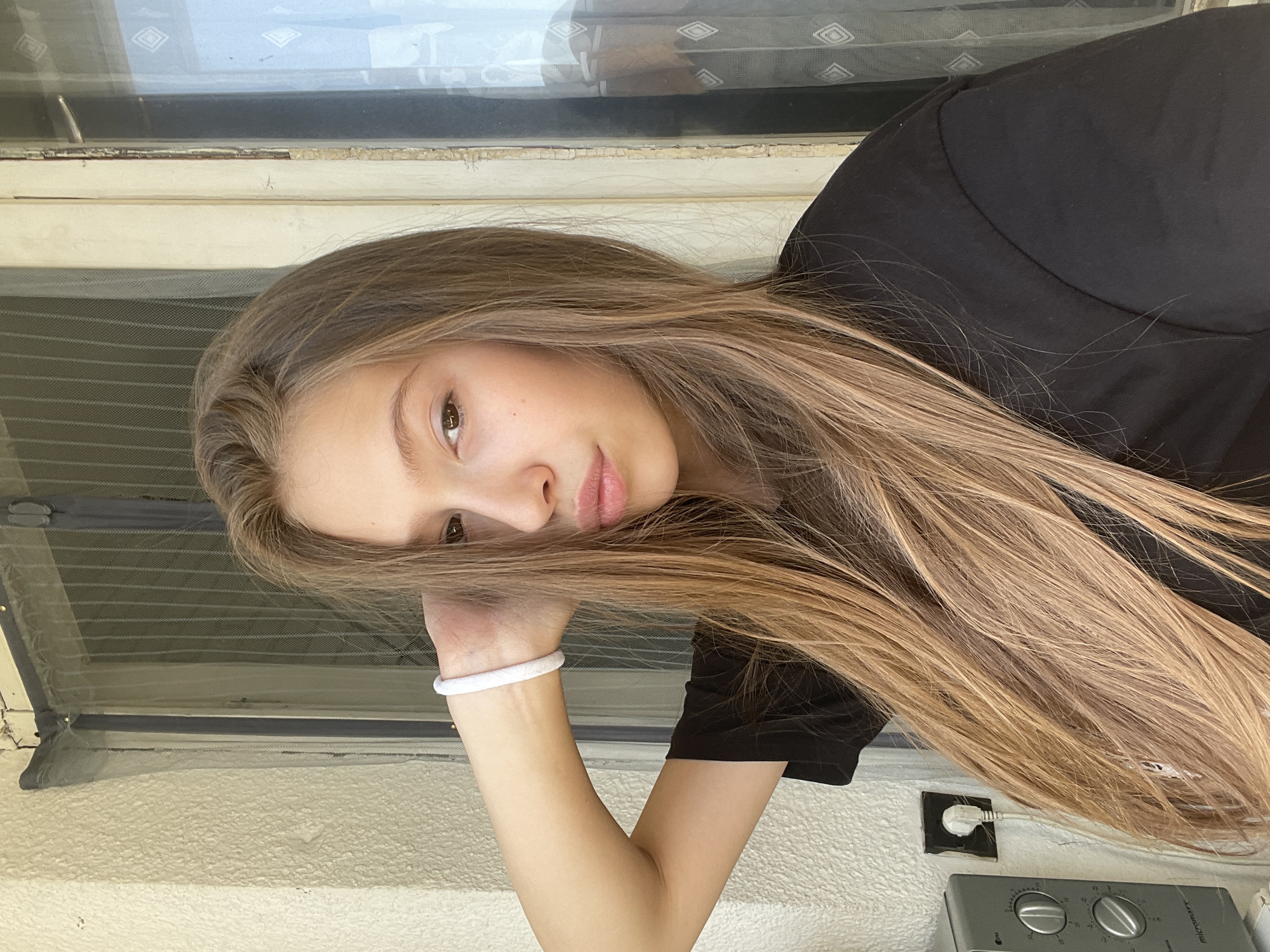 A young woman with long, blonde-brown hair is posing for a selfie outdoors. She is wearing a black t-shirt and a white bracelet on her left wrist, which is raised to her head. Her gaze is directed towards the camera, and her lips are slightly parted. The background consists of a textured wall on the left, a window with a screen, and a door with white trim on the right. A microwave is visible on the wall to the left. The scene appears to be on a balcony or porch. The location is identified as Kyustendil, Bulgaria, based on the provided information.