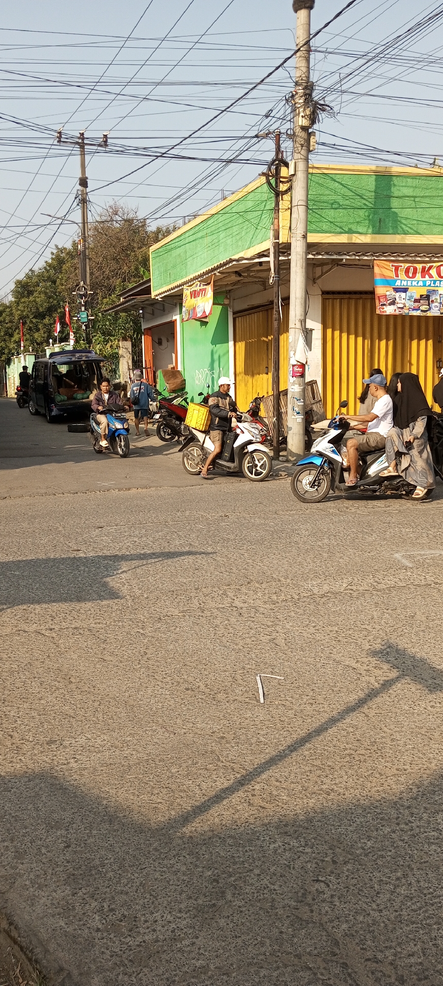 The image depicts a street scene in Setu, Indonesia, bathed in the late afternoon sun, casting long shadows. A busy intersection is captured, with a variety of vehicles and people present. In the foreground, the asphalt road is textured and shows signs of wear. A small white object, possibly a piece of litter or debris, lies on the road.
Several motorcycles are visible, some with riders and some parked. One man in a white cap and shorts is riding a white motorcycle, carrying a yellow crate on the back. Another man, also wearing a white cap, is riding a blue and white motorcycle with a woman in a black hijab and patterned dress as a passenger. A third man in a blue shirt and jeans is walking behind a parked red motorcycle. Further down the road, a blue minivan is parked, its rear door open, revealing cargo inside. A person is visible standing near the open door.
The background features a building with a vibrant green facade and a yellow corrugated metal door. A sign reading "VENTI" is displayed on the building. Another sign above a doorway, partially obscured, appears to advertise "TOKO ANEKA PLASTIK". Utility poles with a dense network of electrical wires crisscross the sky, a common sight in urban Indonesian landscapes. Trees with green foliage are visible in the distance. Two Indonesian flags are planted on the left side of the image. The overall impression is one of everyday activity in a bustling Indonesian town.