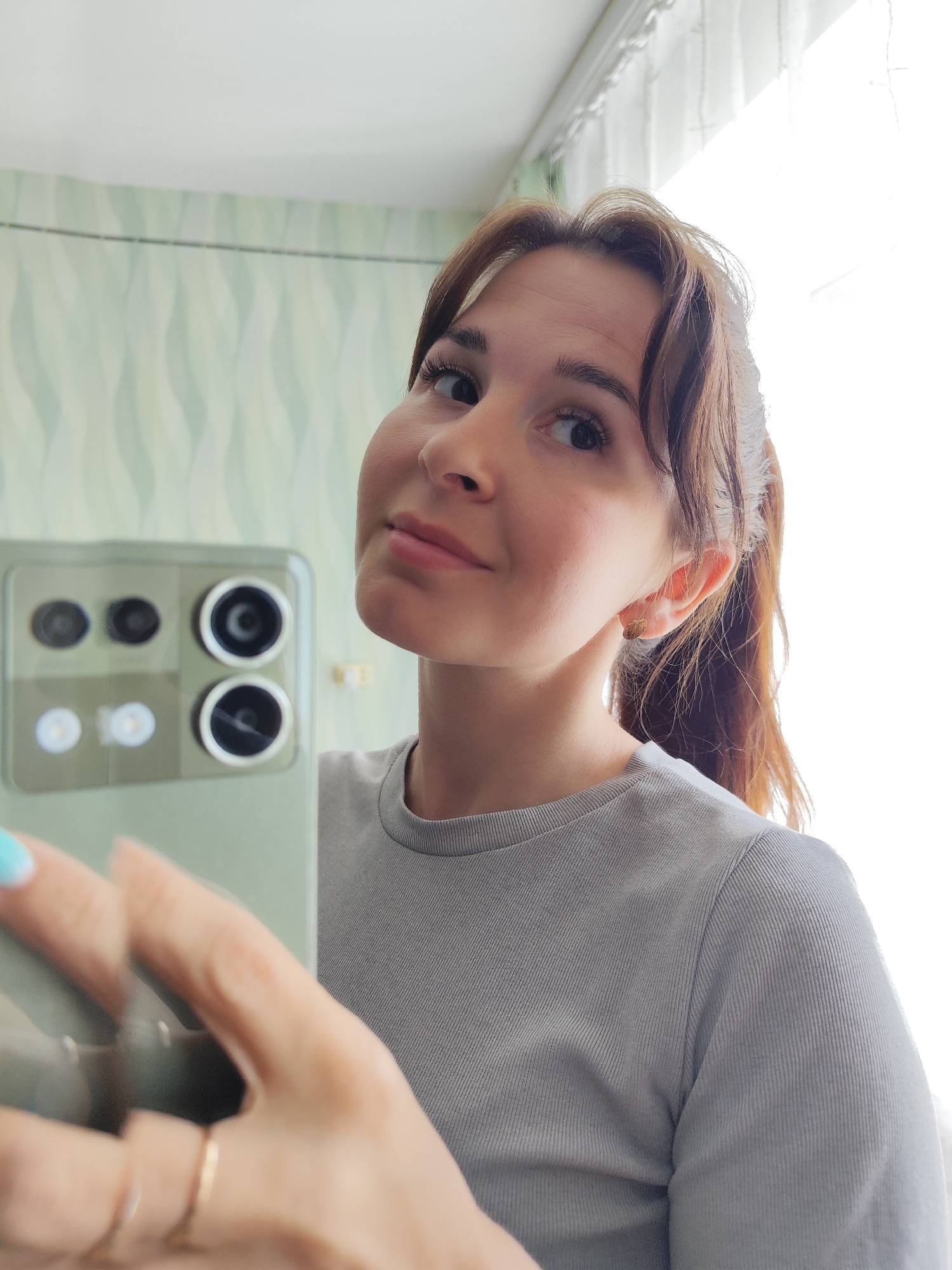 A woman with brown hair pulled back in a ponytail takes a selfie with a smartphone. She is wearing a gray ribbed t-shirt and gold rings on her fingers. The smartphone has multiple camera lenses on the back. The background includes a window with white curtains and a wall with a wavy, light green pattern. The location in the city of Фоки, Russia is not discernible.