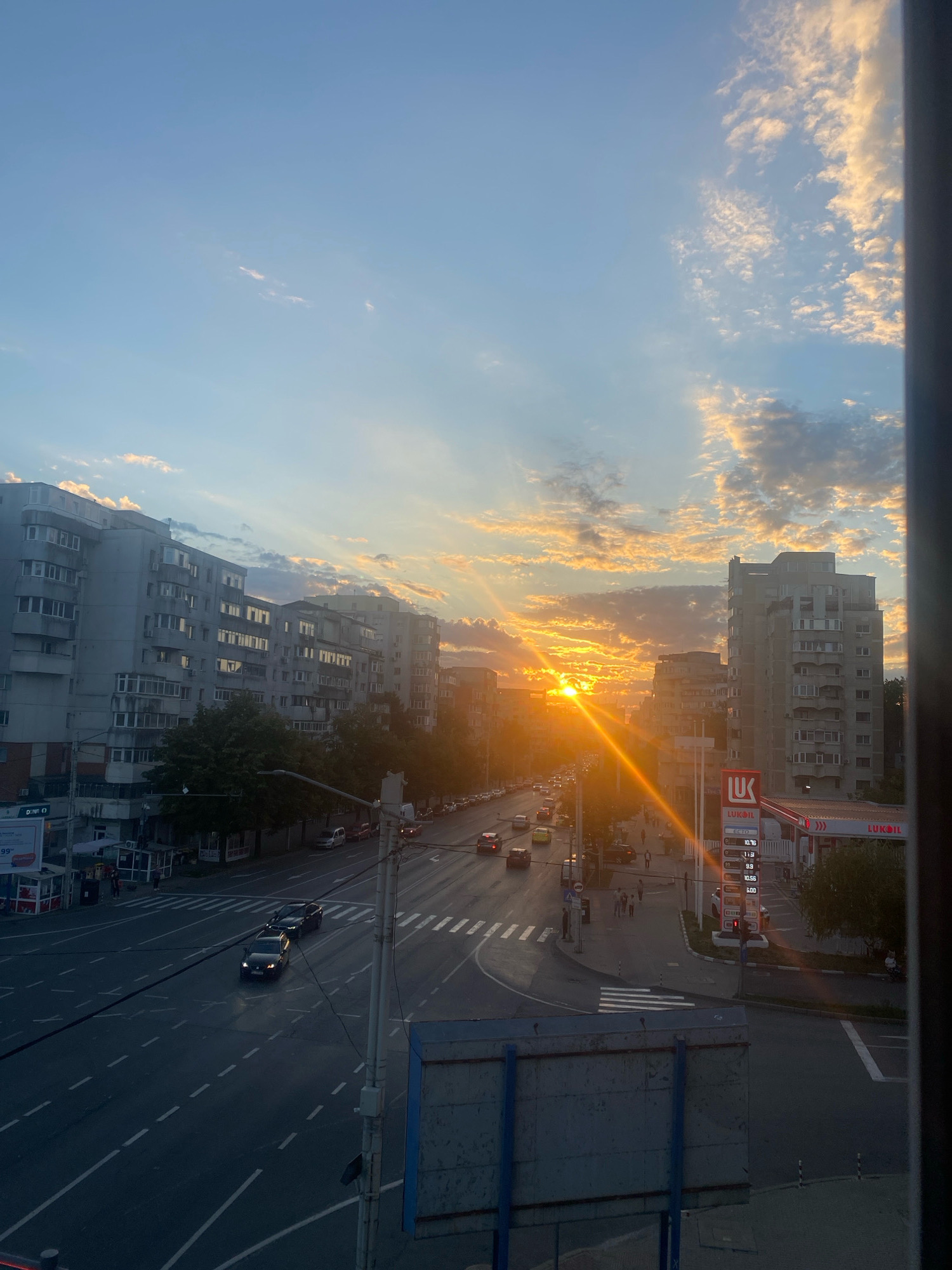 The image captures a street scene in Iasi, Romania, during sunset. The sun is low on the horizon, casting a warm, orange glow and creating visible light rays that pierce through the clouds. The sky is a gradient of blue and orange, with scattered, fluffy clouds illuminated by the setting sun.
In the foreground, a road with multiple lanes is visible, marked with white dashed lines and pedestrian crossings. Several cars are driving on the road, appearing as dark silhouettes against the illuminated sky. People are walking on the sidewalks alongside the road.
Dominating the mid-ground and background are multi-story apartment buildings, typical of urban Romanian architecture. Their facades are a muted gray, and many windows reflect the sunset light. Trees with dense green foliage are interspersed between the buildings.
To the right, a Lukoil gas station is prominent. The station has a red and white canopy, and a digital display shows fuel prices. A sign with the Lukoil logo is clearly visible. The overall impression is a bustling city street at dusk, with the dramatic backdrop of a colorful sunset.