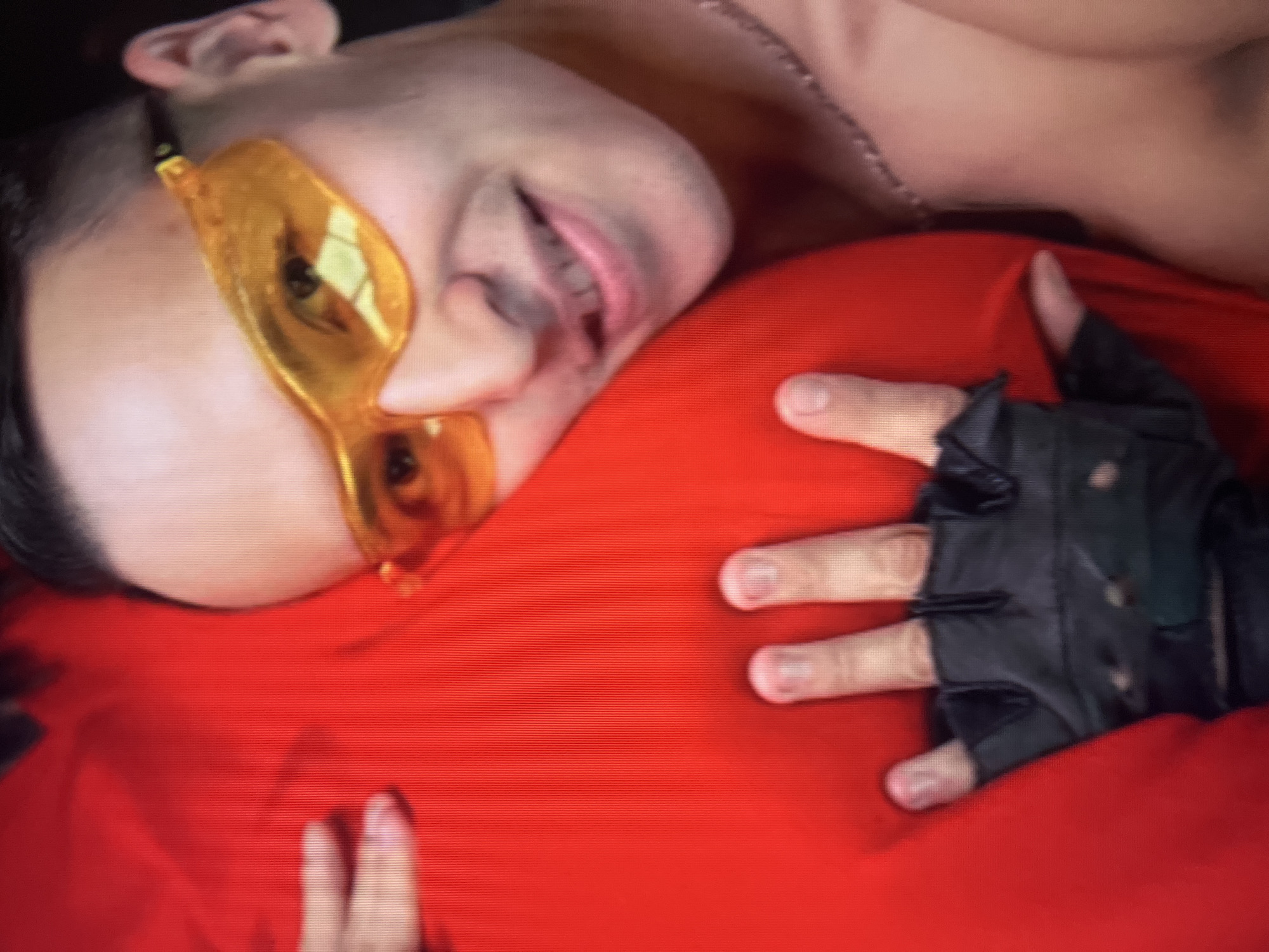 A close-up shot shows a person with short dark hair and yellow sunglasses embracing a large red object. The person's face is partially obscured by the object, and they are smiling with their mouth open. One hand, wearing a black fingerless glove, is visible resting on the red object. The background is dark and indistinct. There is no discernible information about the location being Qarağandı city administration, Kazakhstan.
