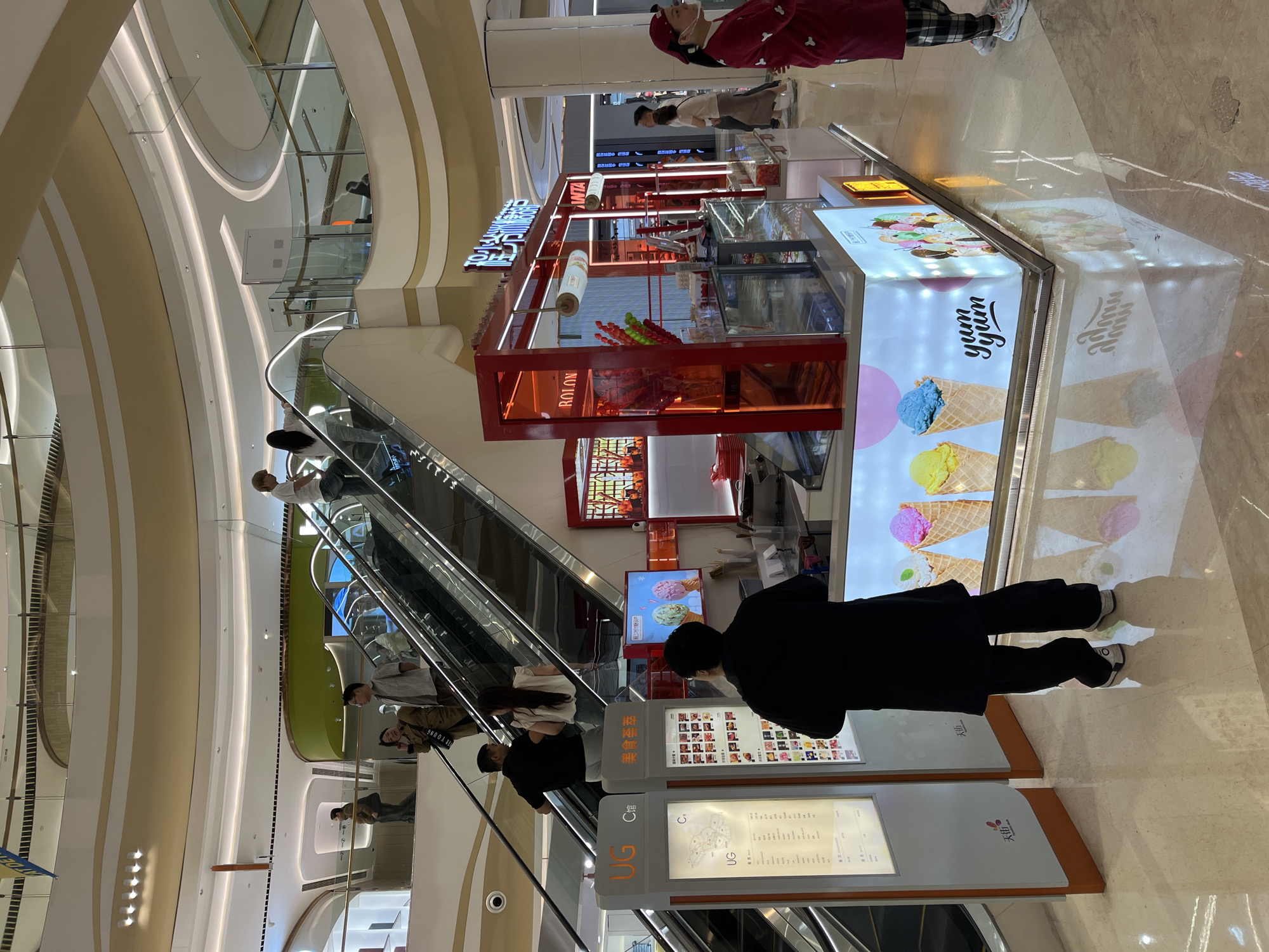 This photograph, taken indoors at a shopping mall, depicts an ice cream stand in Yuzhong District, China. The stand, labeled "Yum Yum" and advertising a variety of ice cream flavors, is positioned near a set of escalators. Several individuals are present in the scene: one person, dressed in black, stands in front of the ice cream stand, appearing to examine a menu displayed on a nearby screen. Other individuals are seen on the escalators, traveling between floors of the mall. The environment includes signage in both English and Chinese, further identifying the location and offerings available at the stand and throughout the mall.