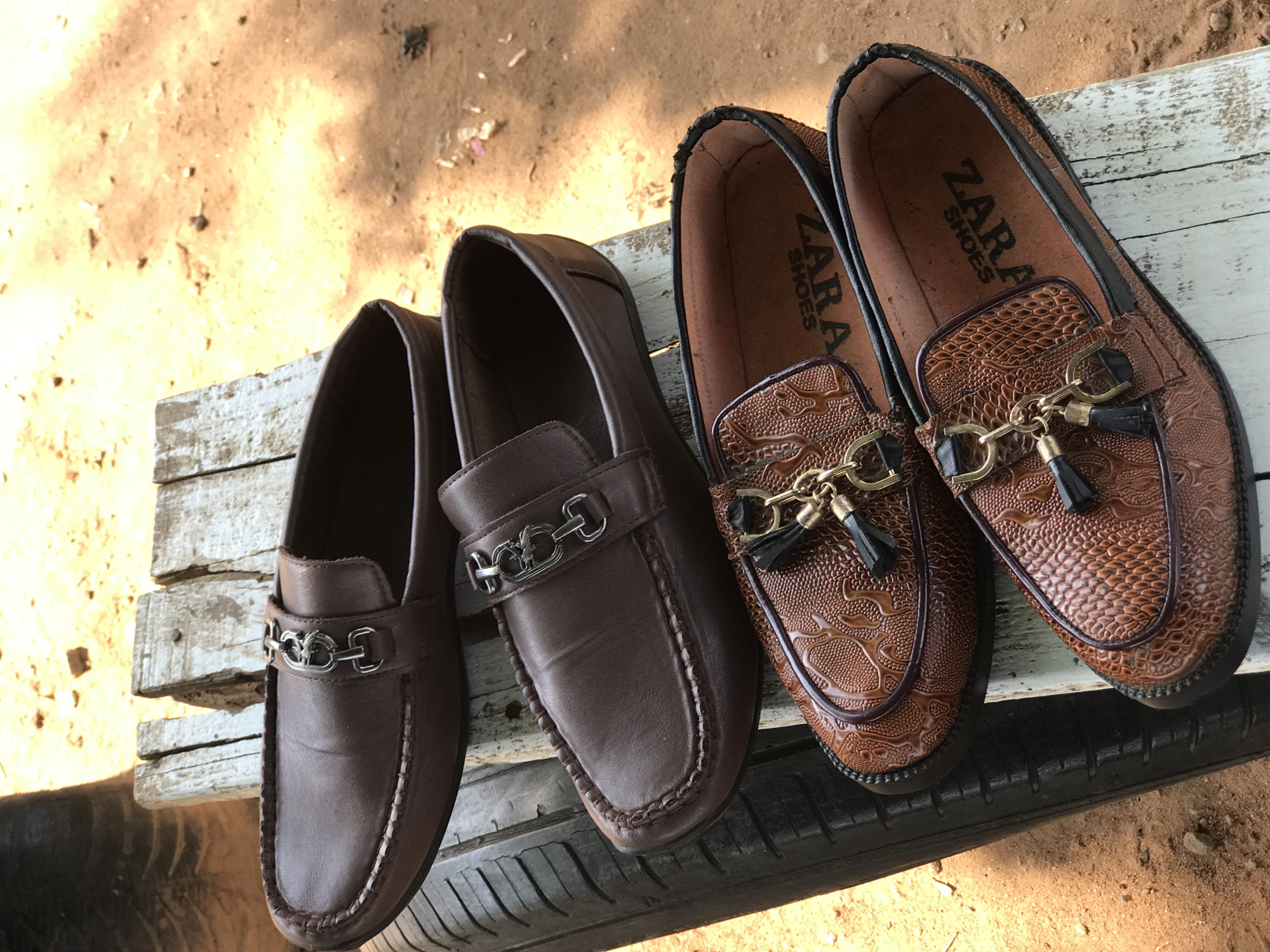 Two pairs of men's loafers are arranged on a weathered wooden surface atop what appears to be a vehicle tire. The first pair is a solid, matte brown leather with a silver-colored chain detail across the vamp. The second pair, likely of the same style, features a textured brown leather, embossed with a reptilian or floral pattern, and adorned with a gold-colored chain and tassel detail. The interior of this second pair reveals "ZARA SHOES" printed inside. The background consists of dry, sandy ground. The image captures a casual setting, possibly an outdoor market or roadside display, given the tire and rough wooden surface. It is impossible to verify if the location is Abuja, Nigeria.