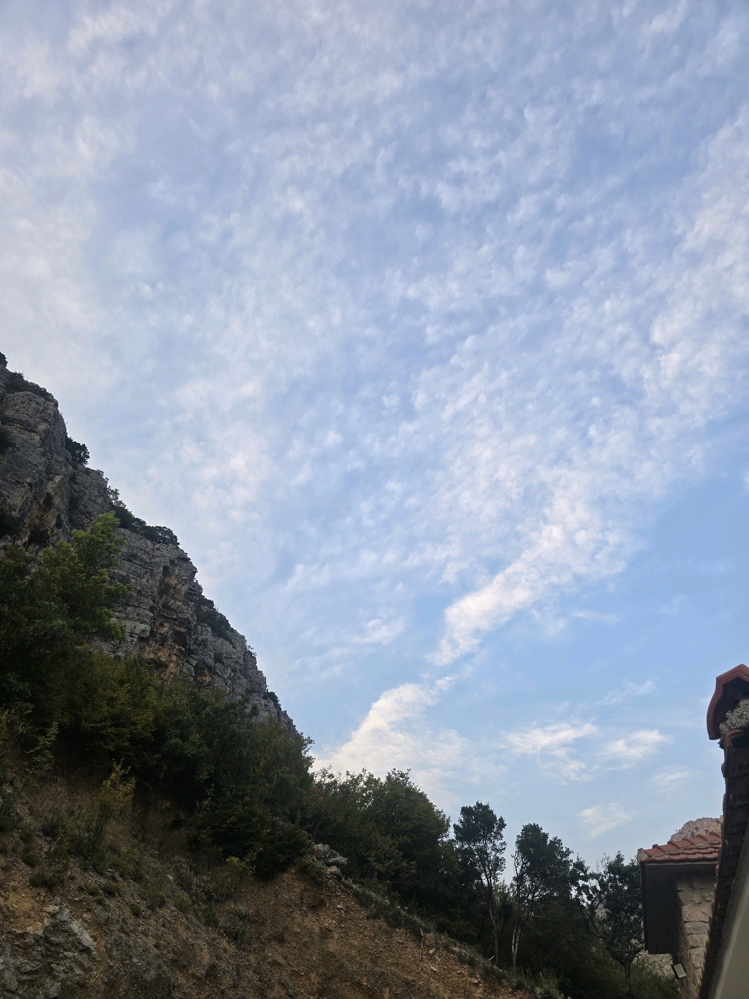 The image depicts a rugged, mountainous landscape with a steep, rocky incline on the left side, covered in dense green foliage. The sky above is a pale blue, streaked with wispy white clouds. In the lower right corner, a portion of a building with a terracotta tiled roof and stone walls is visible. The environment appears to be natural and undeveloped, suggesting a location outside of the immediate urban center, possibly in the hills surrounding Podgorica, Montenegro, where such natural scenery is common. There are no people or animals visible in the frame. The scene is captured in natural daylight, with the lighting suggesting it might be late afternoon or early morning given the soft illumination.
