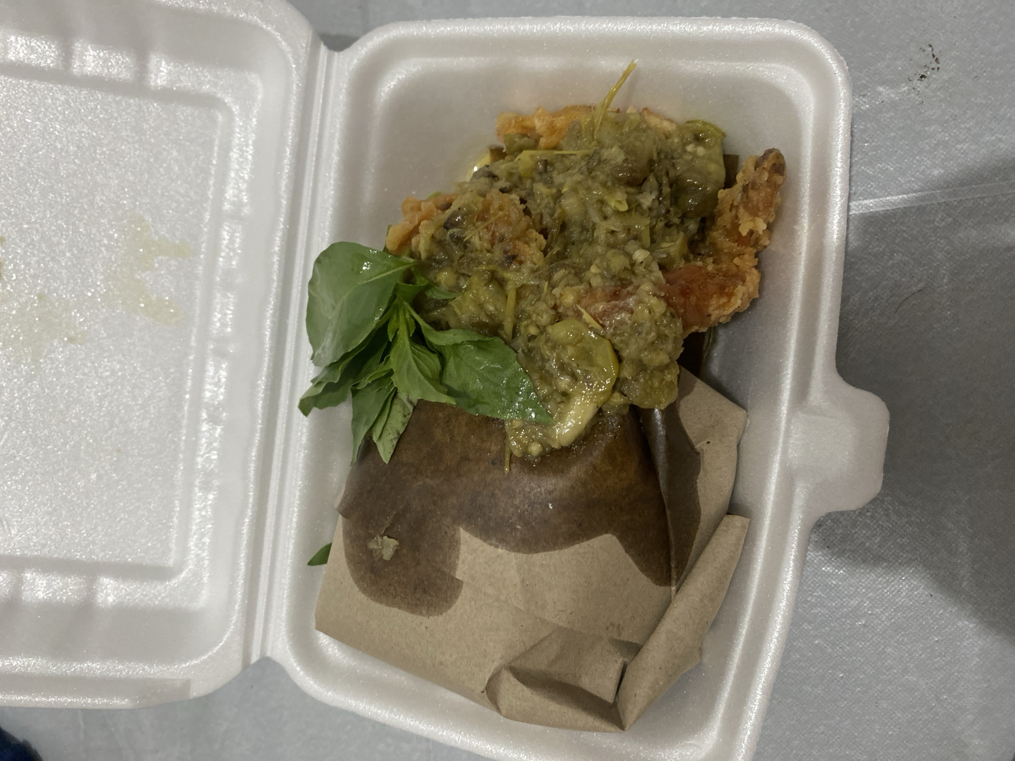 The image shows a close-up, overhead view of a takeout food container. The container is white styrofoam and appears to be open. Inside, the food is placed on brown paper. The food consists of what appears to be fried chicken or a similar item, covered in a green chili sauce and garnished with fresh basil leaves. There is a visible liquid sheen on the food and within the container, suggesting it may be oily or saucy. The background is out of focus but appears to be a textured surface, possibly a table. The lighting suggests an indoor setting, potentially a home or restaurant environment. No people or specific city landmarks are visible. The style of food packaging suggests it was purchased for takeout or delivery.