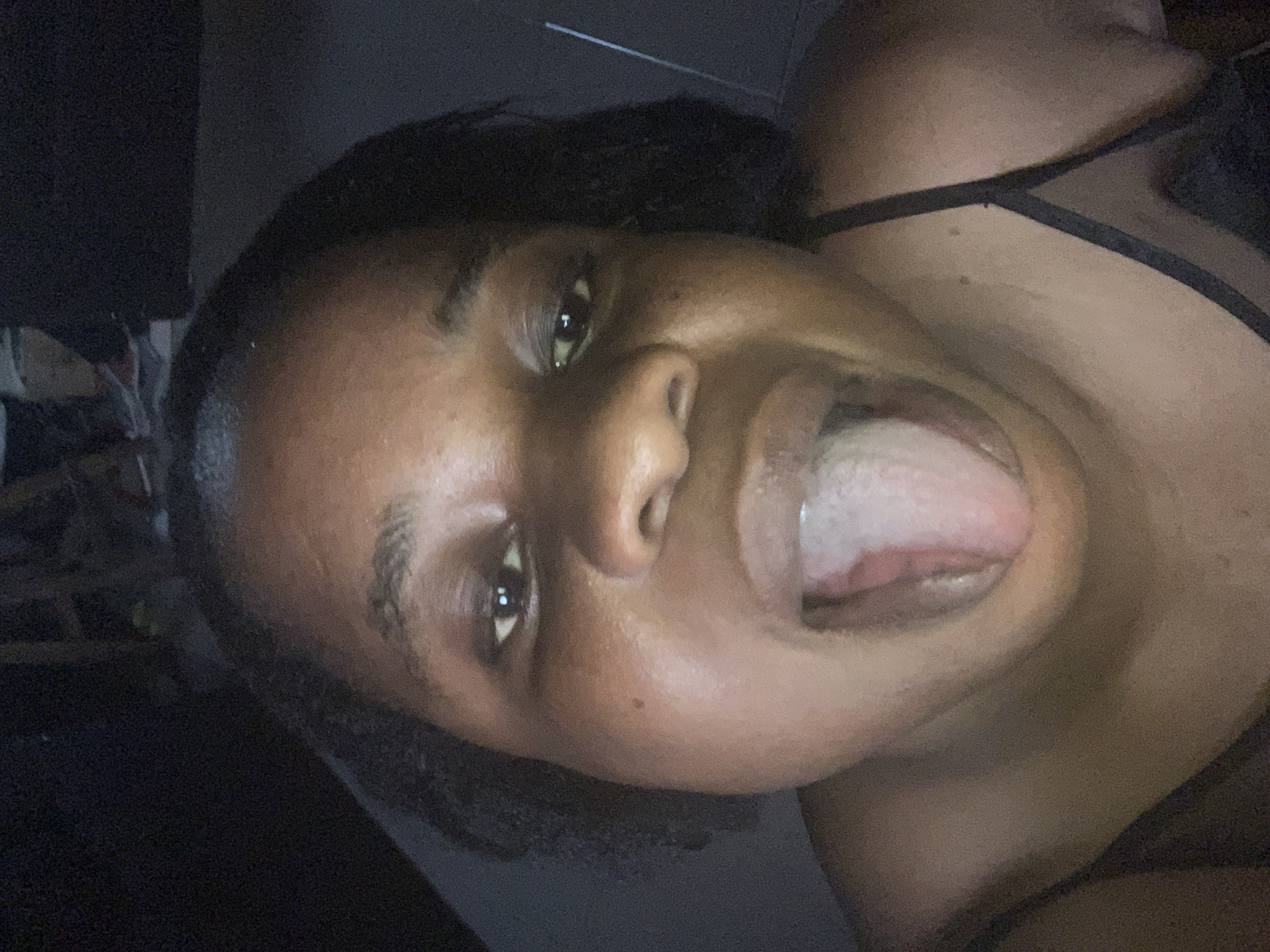 A close-up, slightly angled portrait shows a dark-skinned person with their mouth open and tongue extended. The person has dark hair, and a black strap is visible along the upper right side of the image, suggesting clothing or undergarments. The background is dark and indistinct, with some blurred shapes visible in the upper left corner. The location is not specified.