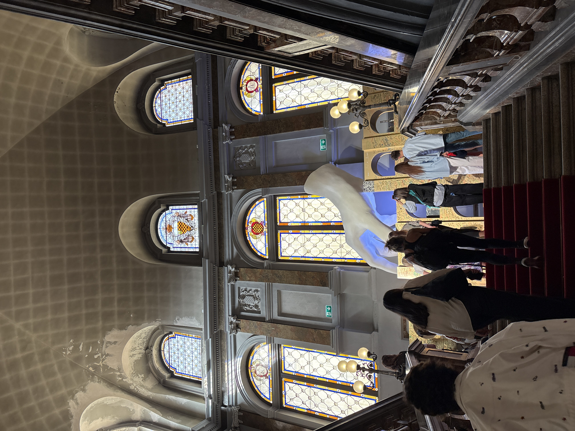 The image depicts an interior shot of a building in Milan, Italy, specifically the Fondazione Prada. The location is recognizable by the white serpentine sculpture visible against the stained-glass windows, and the unique architecture of the stairwell and ceiling.
People are ascending a red-carpeted staircase. The architectural features include arched stained-glass windows, ornate molding, a patterned ceiling, and decorative lighting fixtures. The walls adjacent to the stairs are adorned with what appear to be displays containing writing and QR codes.