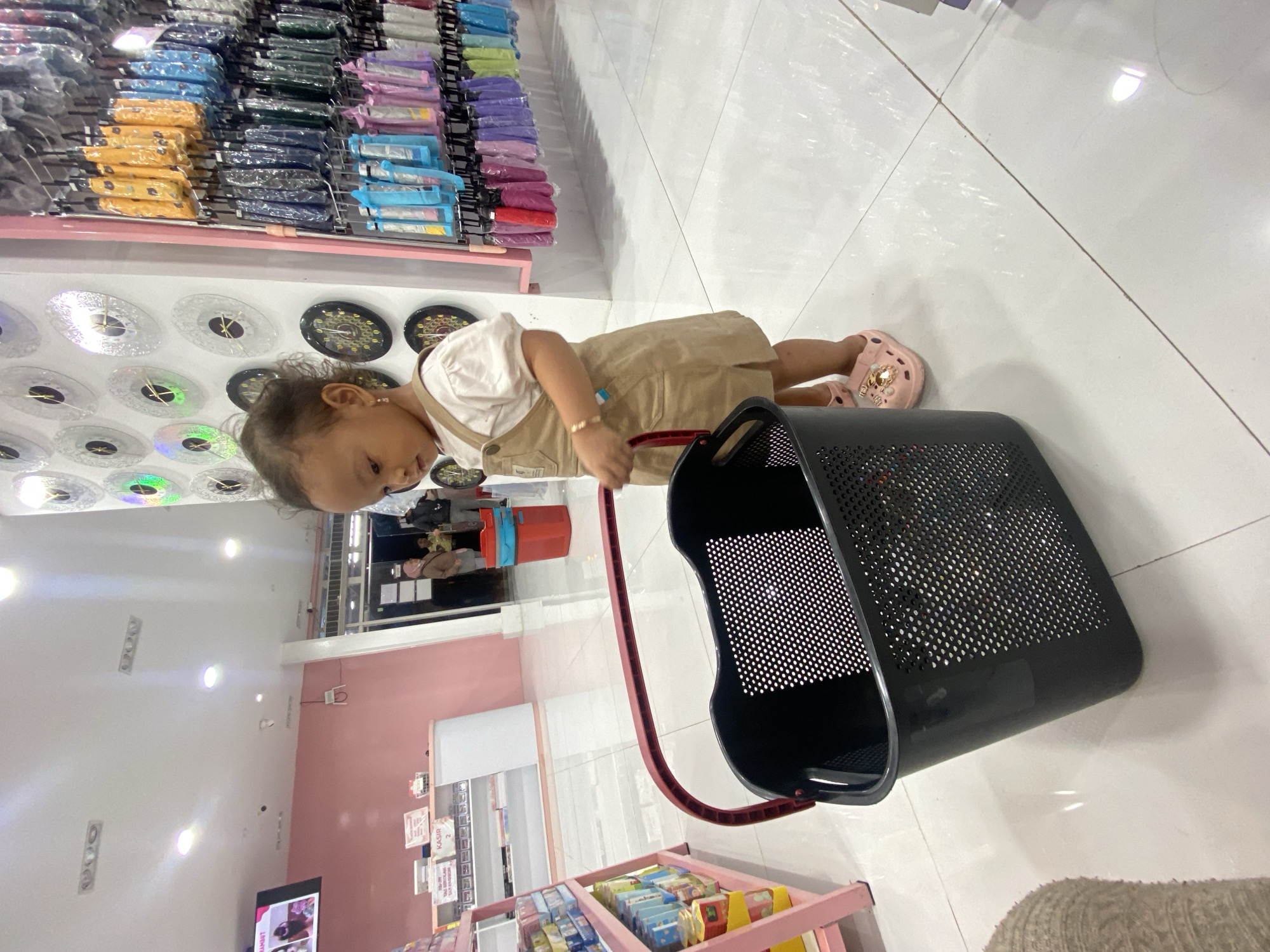 In a well-lit retail store, a young child, likely a toddler, stands facing a black plastic shopping basket. The child wears a light-colored t-shirt and a brown overall dress, with pink clogs on their feet. The basket, with a red handle, is positioned in the foreground. In the background, shelves stocked with various items, including clocks and umbrellas, are visible. The store appears to be in Kecamatan Pati, Indonesia, suggested by the presence of the word "KASIR" (cashier in Indonesian) on a sign in the background. The floor is tiled and reflective. Other shoppers and store employees are visible in the middle ground, indicating an active commercial environment.