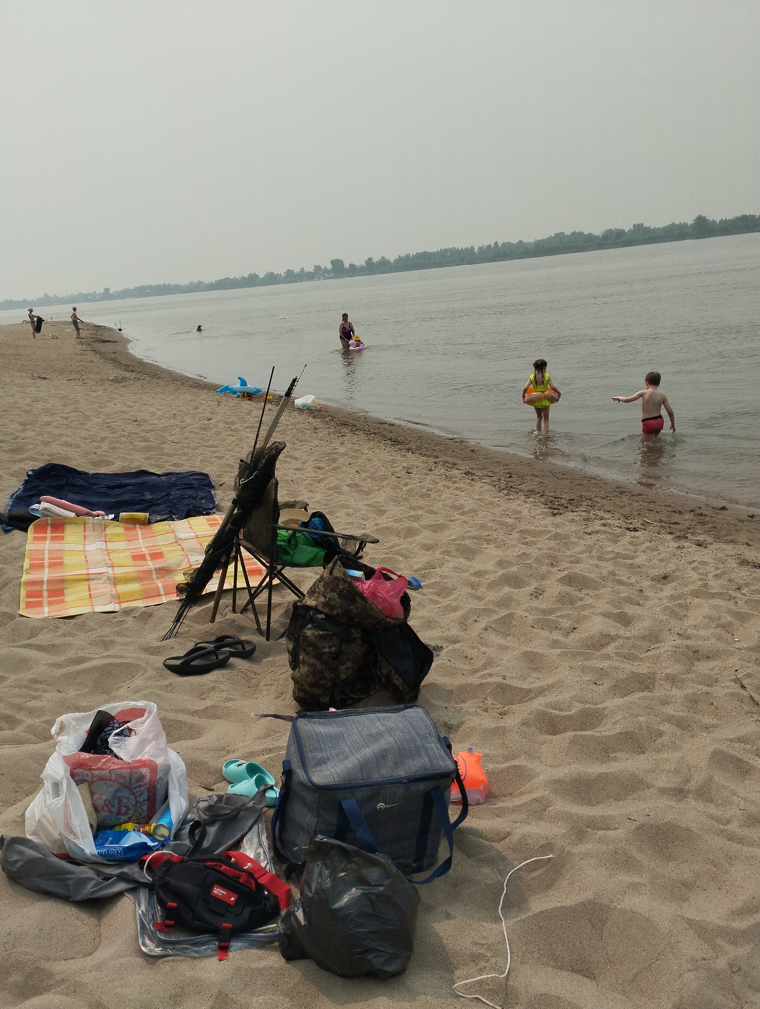 The image depicts a sandy beach with a body of water in the background. Several individuals are present in the water, including two children, one wearing a yellow life vest and the other in red shorts. An adult woman in a purple swimsuit is also in the water, near a pink flotation device. On the beach, a variety of items are laid out, suggesting a picnic or outing. These include beach towels, a blue net, a folding chair with fishing rods, a camouflage bag, a blue cooler, several plastic bags containing items, a black trash bag, and a pair of flip-flops. The sky is overcast, and a tree line is visible in the distance across the water. The scene appears to be a recreational area, likely a beach on the Ob River in Bibikha, Russia, given the context provided.