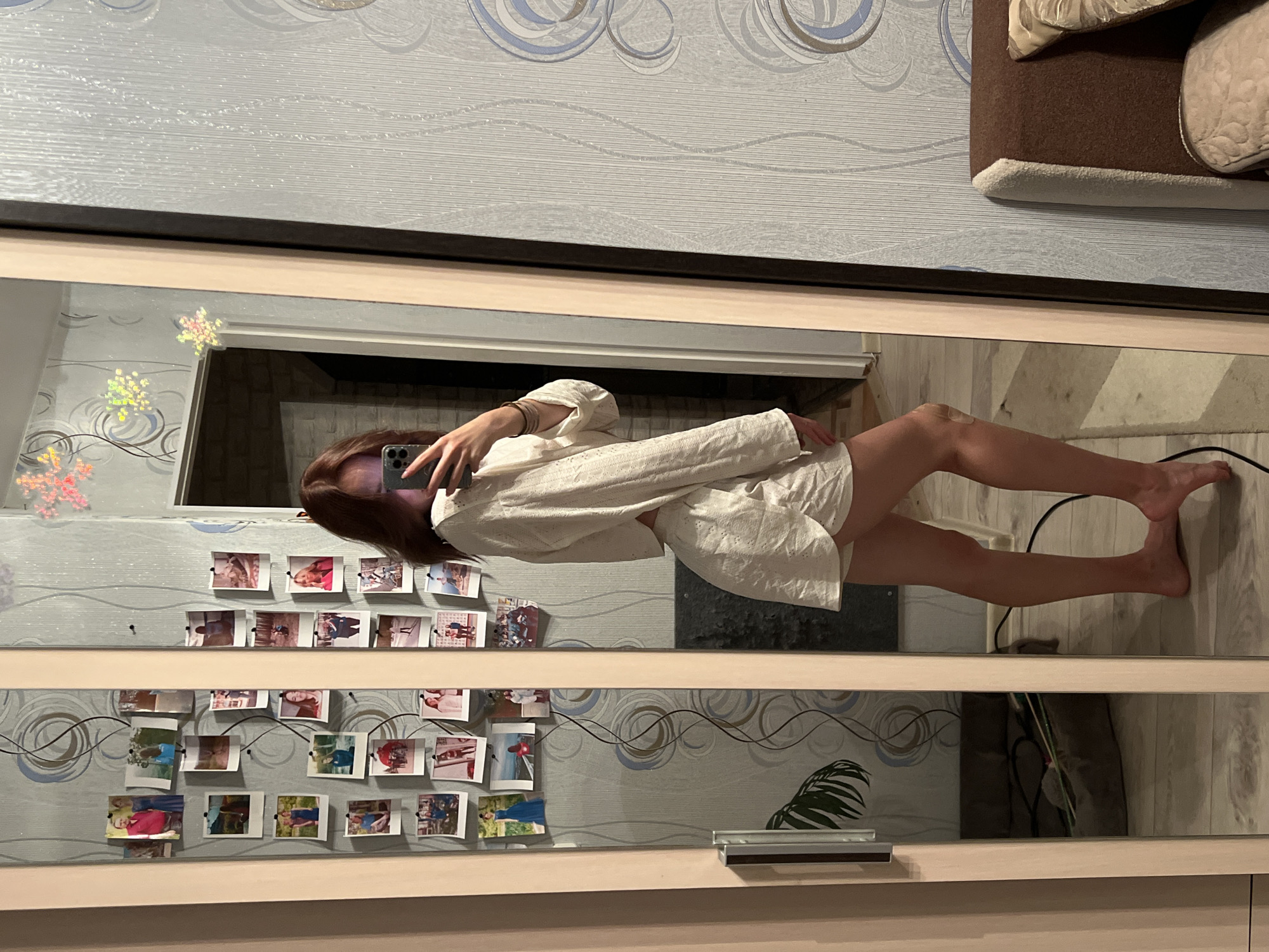 A person is standing in front of a mirror, taking a selfie. The person has medium-length brown hair and is wearing a white top and shorts. The mirror is mounted on a wall, and there are several photographs attached to the wall around the mirror. The wall has patterned wallpaper. The mirror reflects part of the room, including what appears to be a couch and a wooden floor. There's also a plant visible in the reflection. The location is likely indoors, possibly a home in Chaikovsky, Russia.