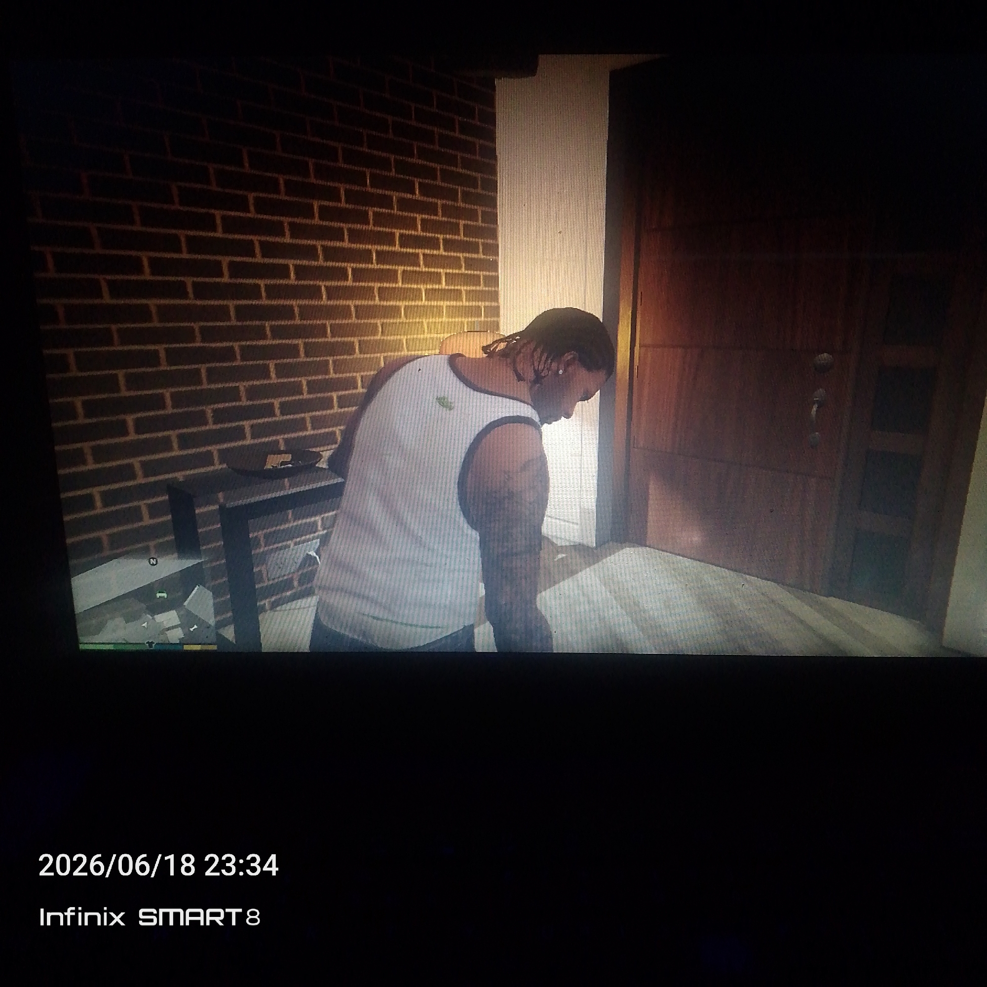 The image depicts a low-angle, full-shot of a person standing in what appears to be an indoor hallway. The individual, who is facing away from the camera and looking downwards, is wearing a white tank top with a dark trim and has dreadlocks. They have tattoos on their arms and are wearing earrings. To the left of the person is a dark, rectangular table with items on it, and beyond that, a brick wall. To the right of the person is a wooden door with panels and a handle, and beyond that, a lighter colored wall. The floor in the hallway appears to be made of wooden planks. A date and time stamp "2026/06/18 23:34" and "Infinix SMART 8" are visible in the bottom left corner of the image, indicating it was taken with an Infinix SMART 8 phone. There are no clear indications of the specific location within a city or any interaction between subjects.