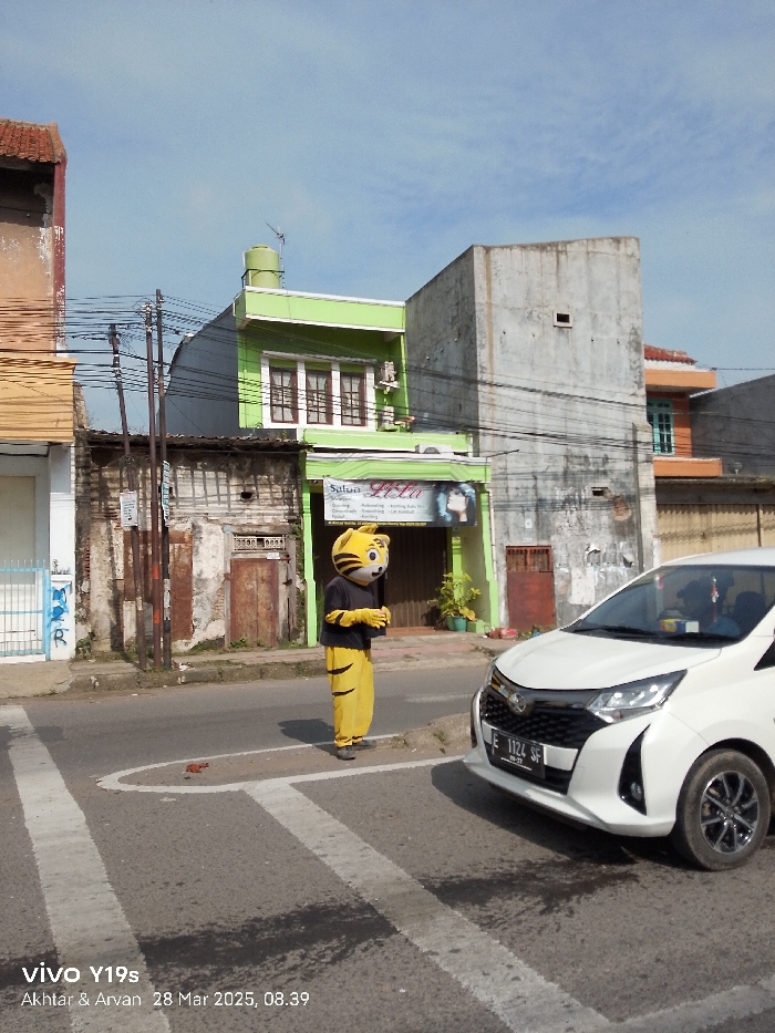 The image shows a person in a yellow and orange tiger costume standing in the middle of a street intersection in Jatibarang, Indonesia. The person is facing the camera, with their arms crossed. The street is paved with asphalt and has white markings for a crosswalk. There are buildings on both sides of the street. There are power lines running across the street and over the buildings. A white Toyota Avanza car is parked on the right side of the image. The car is facing the camera, and its license plate is partially visible. The license plate reads "E 1124 SF." The sky is blue with some white clouds. The image was taken on March 28, 2025, at 08:39 AM.