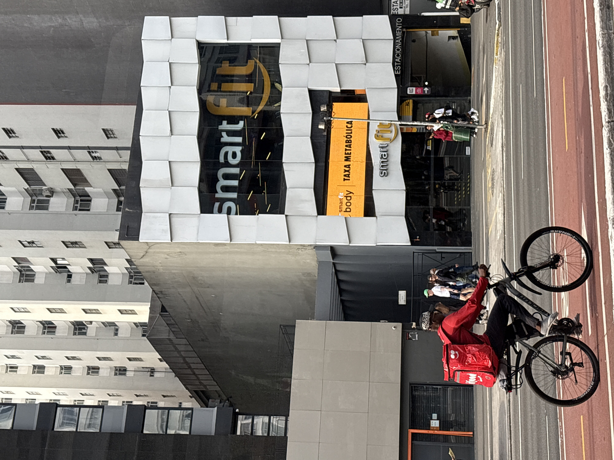 The image captures a street scene in São Paulo, Brazil, specifically in front of a Smart Fit gym. In the foreground, a man on a bicycle rides on a red bike lane. He is wearing a red long-sleeved shirt and black pants, and a large red backpack with a smiley face graphic is strapped to his back. The bicycle is dark gray with silver rims and black tires.
Behind the cyclist, a modern building with a distinctive white, patterned facade houses the Smart Fit gym. The gym's signage, including "smart fit" and a yellow smiley face logo, is prominently displayed on the building. Below the main gym signage, there is an orange sign that reads "smart body TAXA METABÓLICA". To the right of the gym entrance, there is a sign indicating "ESTACIONAMENTO" (parking) with an arrow pointing towards an underground parking garage.
The background features tall, multi-story buildings, characteristic of a dense urban environment. Several people are visible walking on the sidewalk in front of the gym, and other buildings line the street. The overall impression is a bustling city environment with everyday street life and commercial activity.