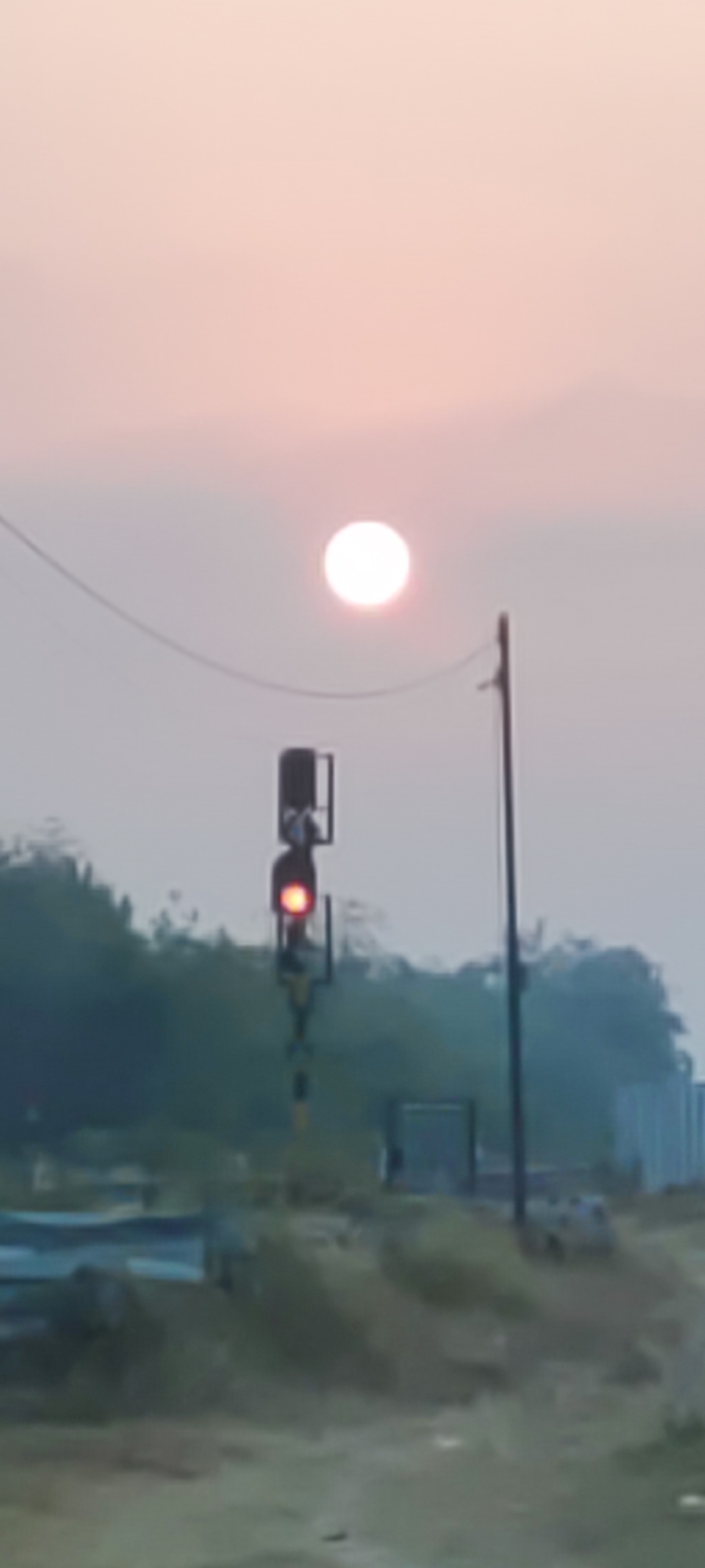 The image shows a railway signal post with a red light illuminated, positioned in front of a hazy, pale sky with a large, soft-edged circle of light, likely the sun partially obscured by atmospheric conditions. The signal post is black and yellow striped at the bottom and features multiple signal lights. Behind the signal, a line of trees and vegetation is visible, with some structures in the background, including what appears to be a blue object. The ground in the foreground is rough and appears to be a natural landscape with some scattered debris. The overall impression is that of a slightly foggy or hazy day, possibly sunrise or sunset, in a rural or semi-rural area near a railway line in Pakusamben, Indonesia. No people are visible in the image.